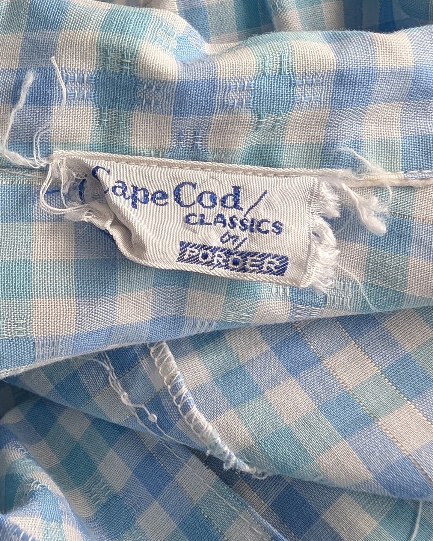 Close-up of a light blue and white gingham fabric with a visible sewn-in label reading Cape Cod Classics by Podor.