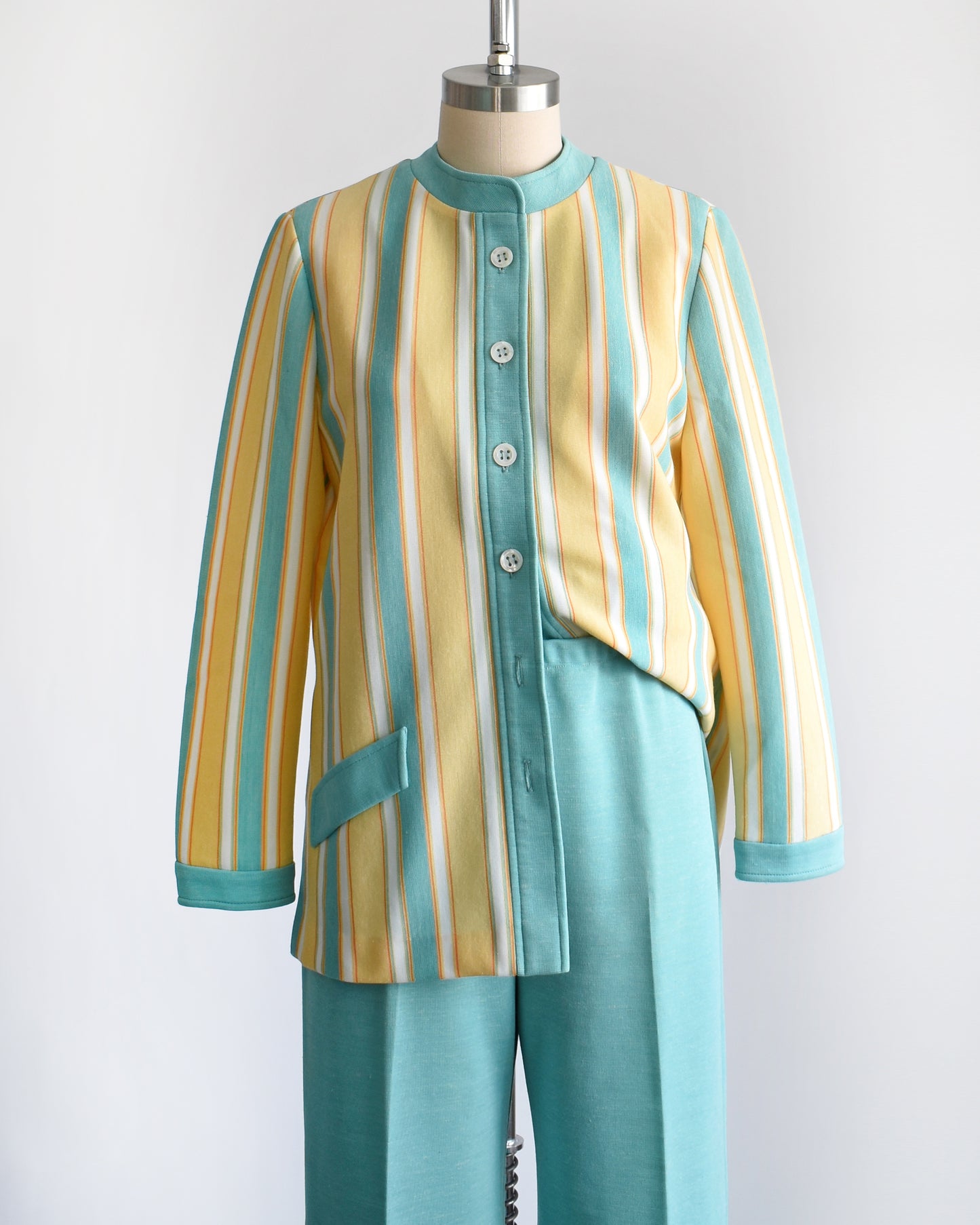 A vintage 1970s two piece pantsuit. The top is yellow, blue, white and orange striped, and the pants are the same matching blue. The top is unbuttoned at the bottom and half tucked into the pants