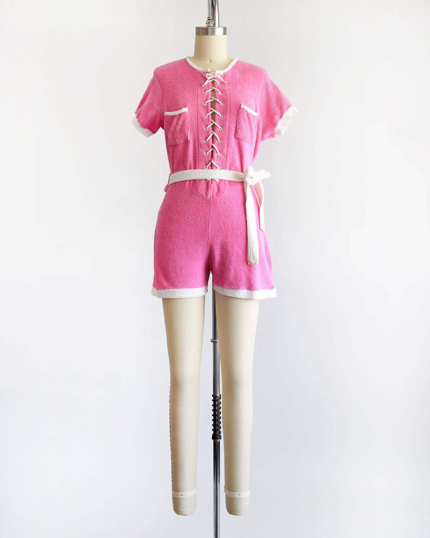 a vintage 1980s pink short-sleeve romper displayed on a cream mannequin against a plain white background. The romper features white trim along the collar, sleeve cuffs, pocket edges, and hem, with a white lace-up front detail and a matching white tie belt at the waist.