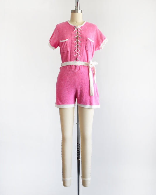 a vintage 1980s pink short-sleeve romper displayed on a cream mannequin against a plain white background. The romper features white trim along the collar, sleeve cuffs, pocket edges, and hem, with a white lace-up front detail and a matching white tie belt at the waist.