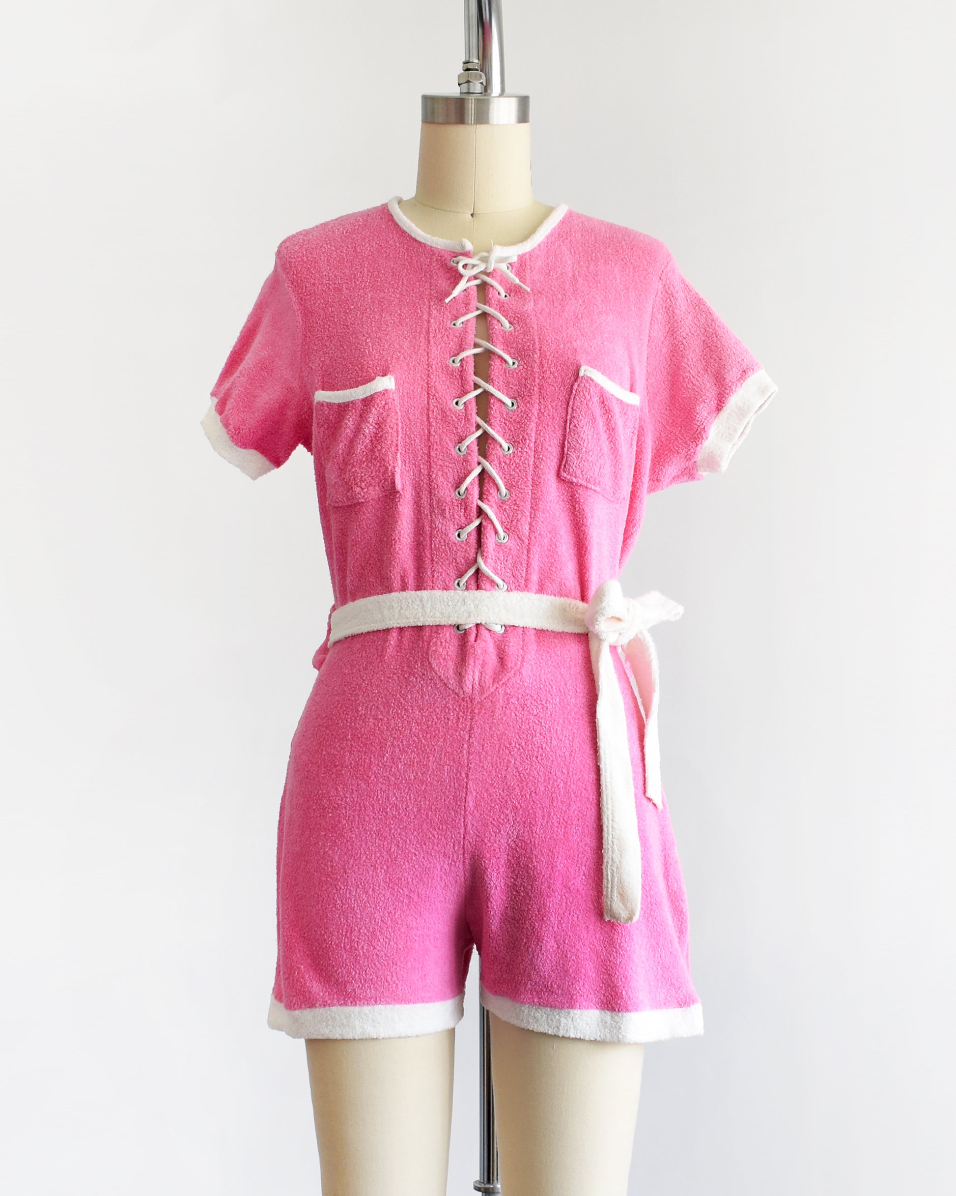 a vintage 1980s pink short-sleeve romper displayed on a cream mannequin against a plain white background. The romper features white trim along the collar, sleeve cuffs, pocket edges, and hem, with a white lace-up front detail and a matching white tie belt at the waist.