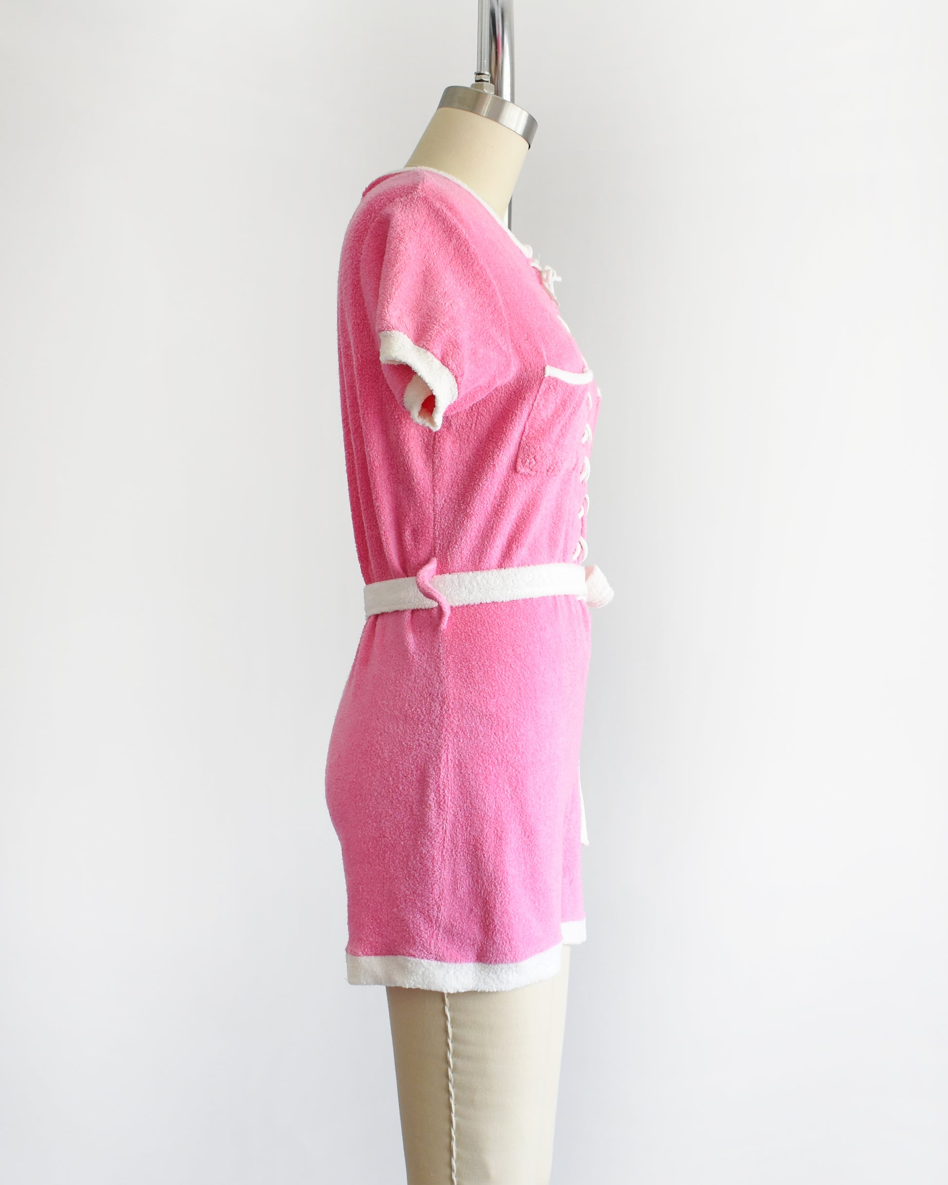 side view of a vintage 1980s pink short-sleeve romper displayed on a cream mannequin against a plain white background. The romper features white trim along the collar, sleeve cuffs, pocket edges, and hem, with a white lace-up front detail and a matching white tie belt at the waist.