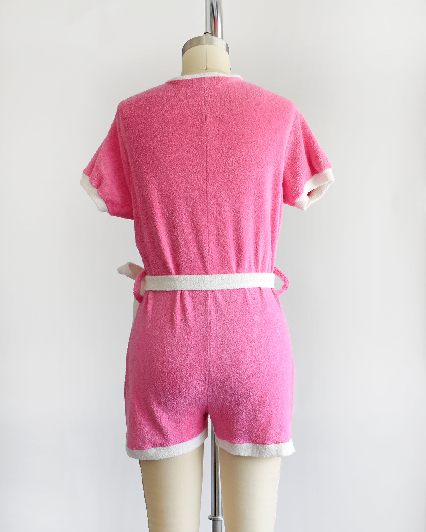 back view of a vintage 1980s pink short-sleeve romper displayed on a cream mannequin against a plain white background. The romper features white trim along the collar, sleeve cuffs, pocket edges, and hem, with a white lace-up front detail and a matching white tie belt at the waist.