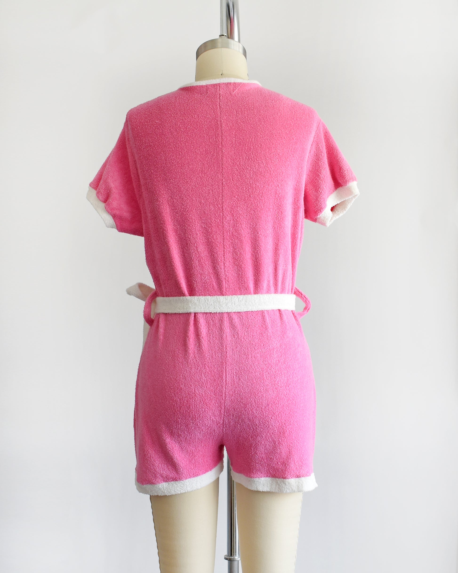 back view of a vintage 1980s pink short-sleeve romper displayed on a cream mannequin against a plain white background. The romper features white trim along the collar, sleeve cuffs, pocket edges, and hem, with a white lace-up front detail and a matching white tie belt at the waist.