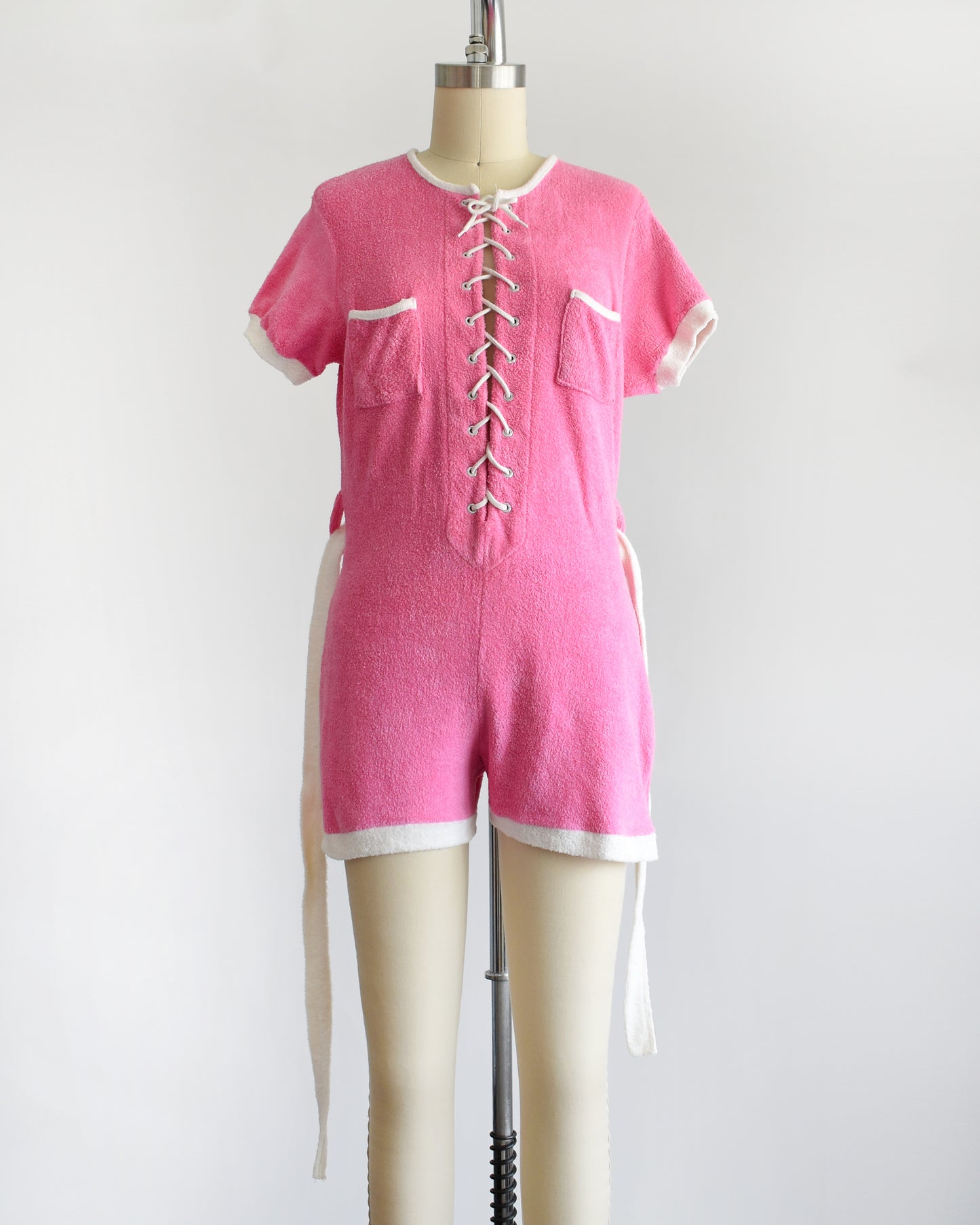 a vintage 1980s pink short-sleeve romper displayed on a cream mannequin against a plain white background. The romper features white trim along the collar, sleeve cuffs, pocket edges, and hem, with a white lace-up front detail and a matching white tie belt at the waist. the belt is undone and hanging on the sides in this photo.