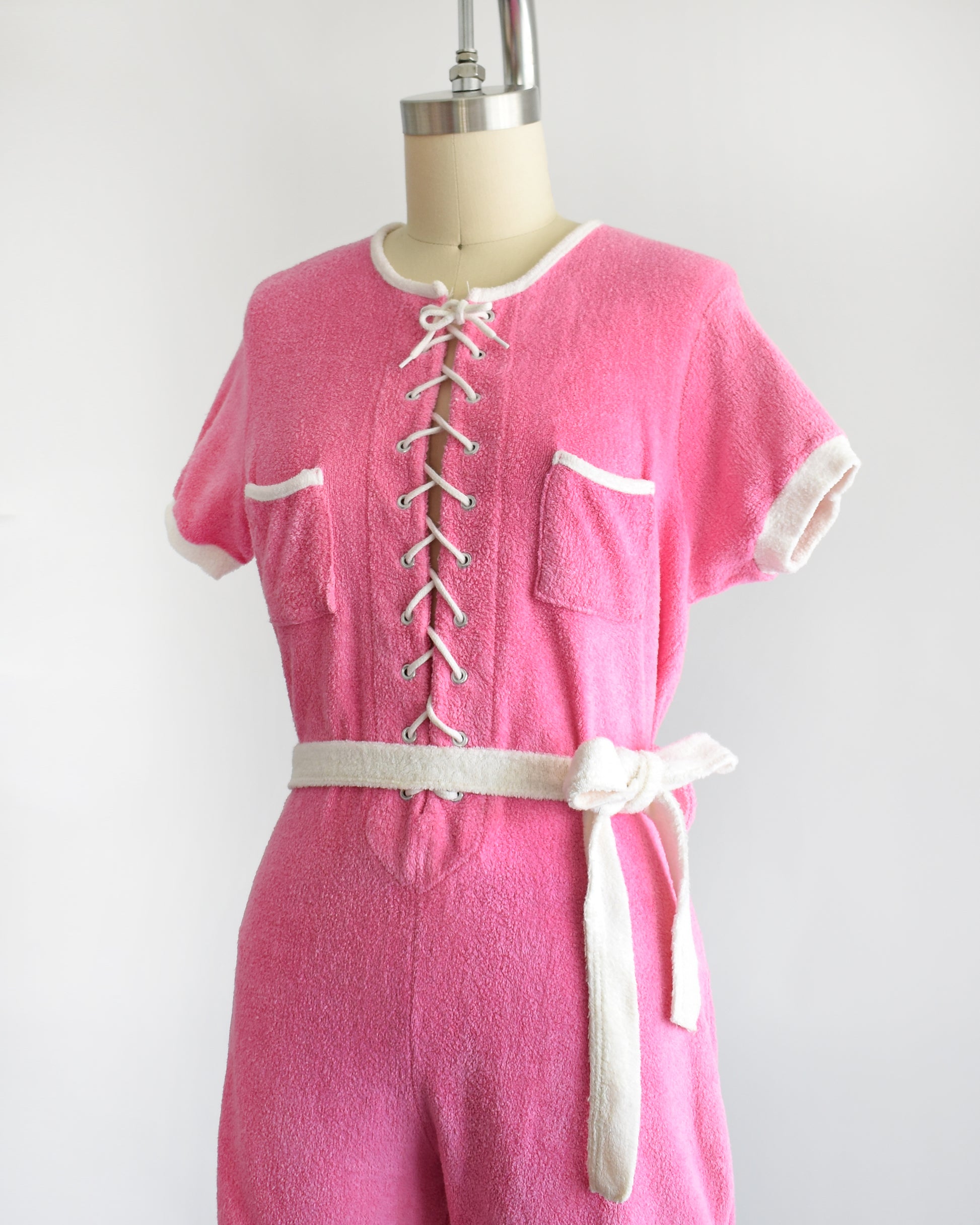 side view of a vintage 1980s pink short-sleeve romper displayed on a cream mannequin against a plain white background. The romper features white trim along the collar, sleeve cuffs, pocket edges, and hem, with a white lace-up front detail and a matching white tie belt at the waist.