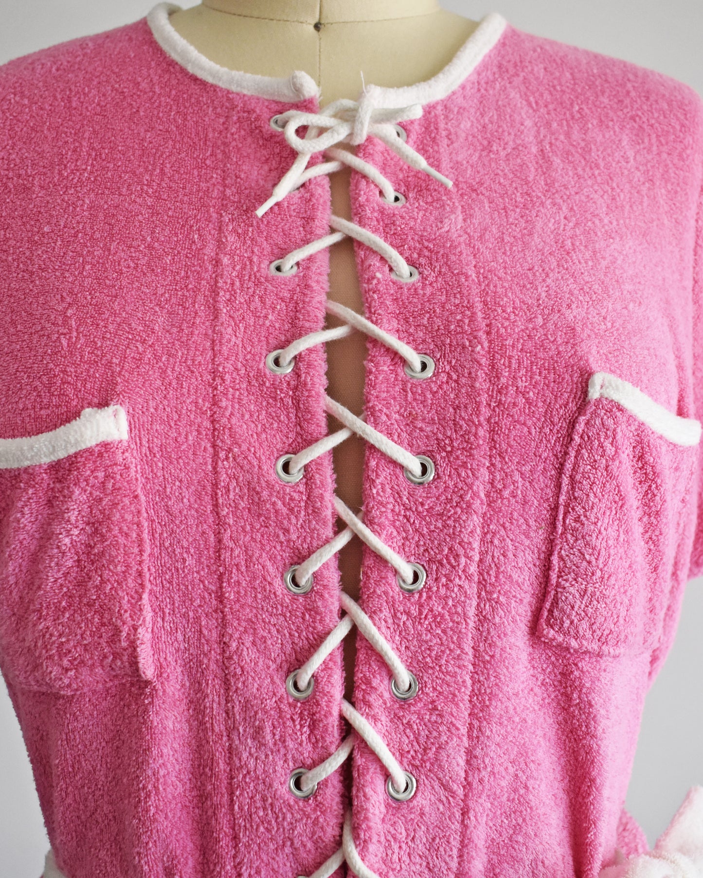 close up of the lace up front on a vintage 1980s pink short-sleeve romper displayed on a cream mannequin against a plain white background. The romper features white trim along the collar, sleeve cuffs, pocket edges, and hem, with a white lace-up front detail and a matching white tie belt at the waist.