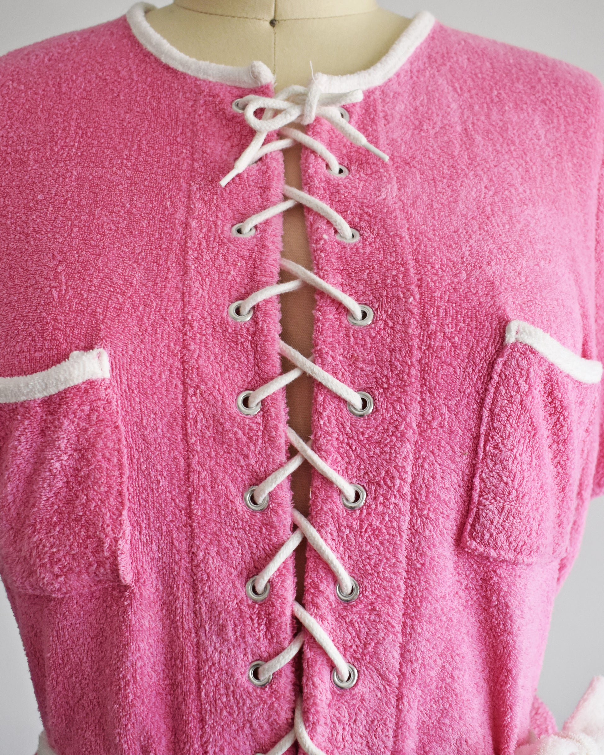 close up of the lace up front on a vintage 1980s pink short-sleeve romper displayed on a cream mannequin against a plain white background. The romper features white trim along the collar, sleeve cuffs, pocket edges, and hem, with a white lace-up front detail and a matching white tie belt at the waist.