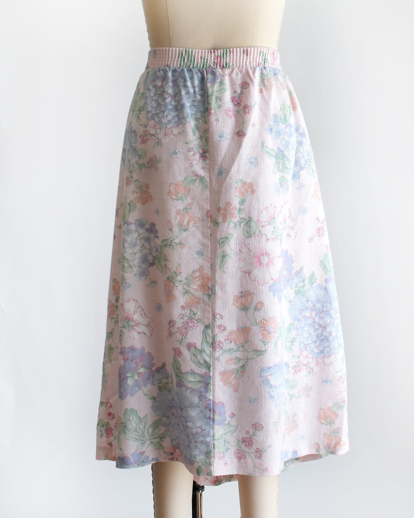 back view of a vintage 1980s  high-waisted midi skirt with a pastel floral print, displayed on a beige dress form mannequin against a plain white background.