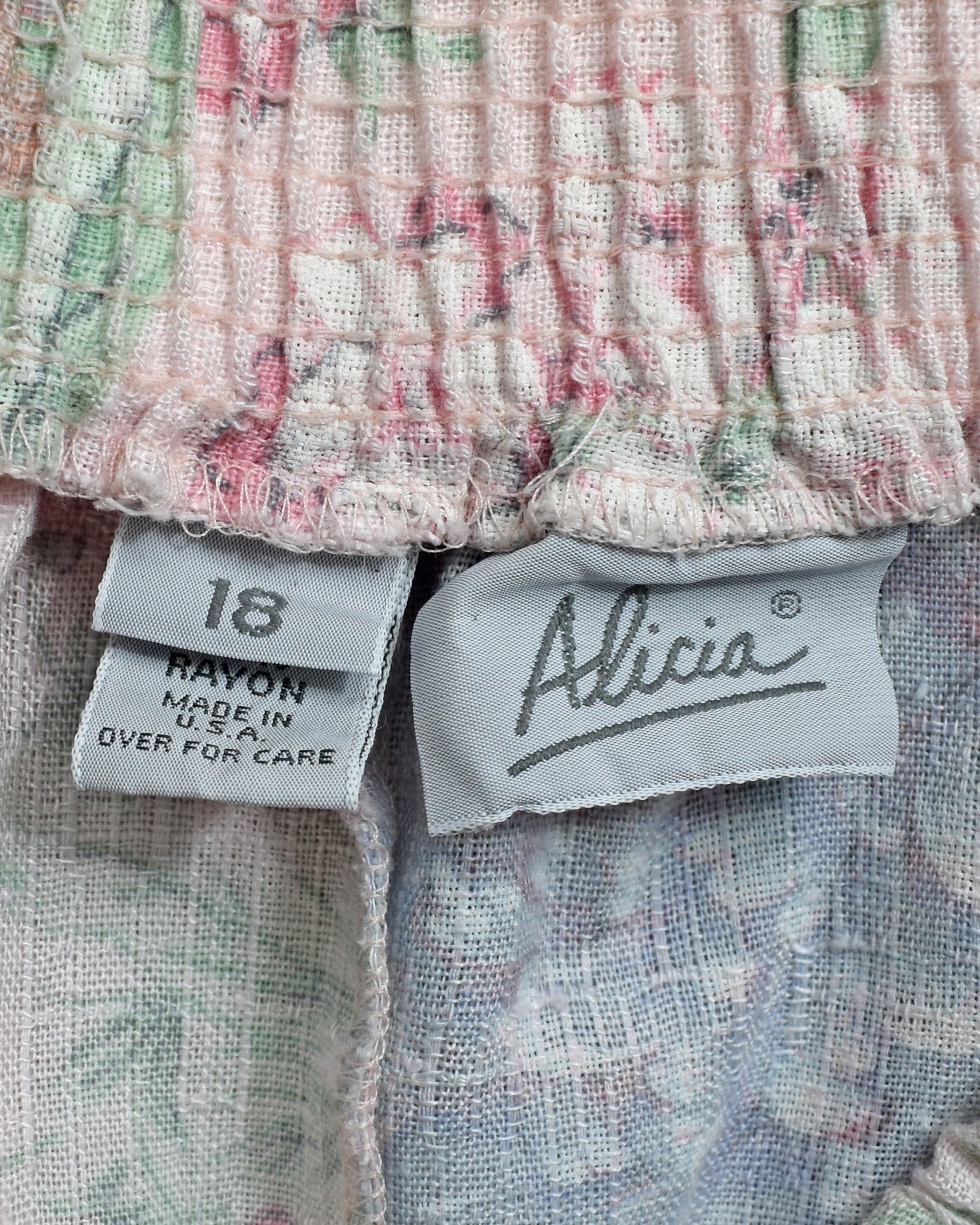 Close-up of a garment's elastic waistband and interior labels, showing a textured woven fabric with a soft pastel multicolor pattern and a visible Alicia brand tag.