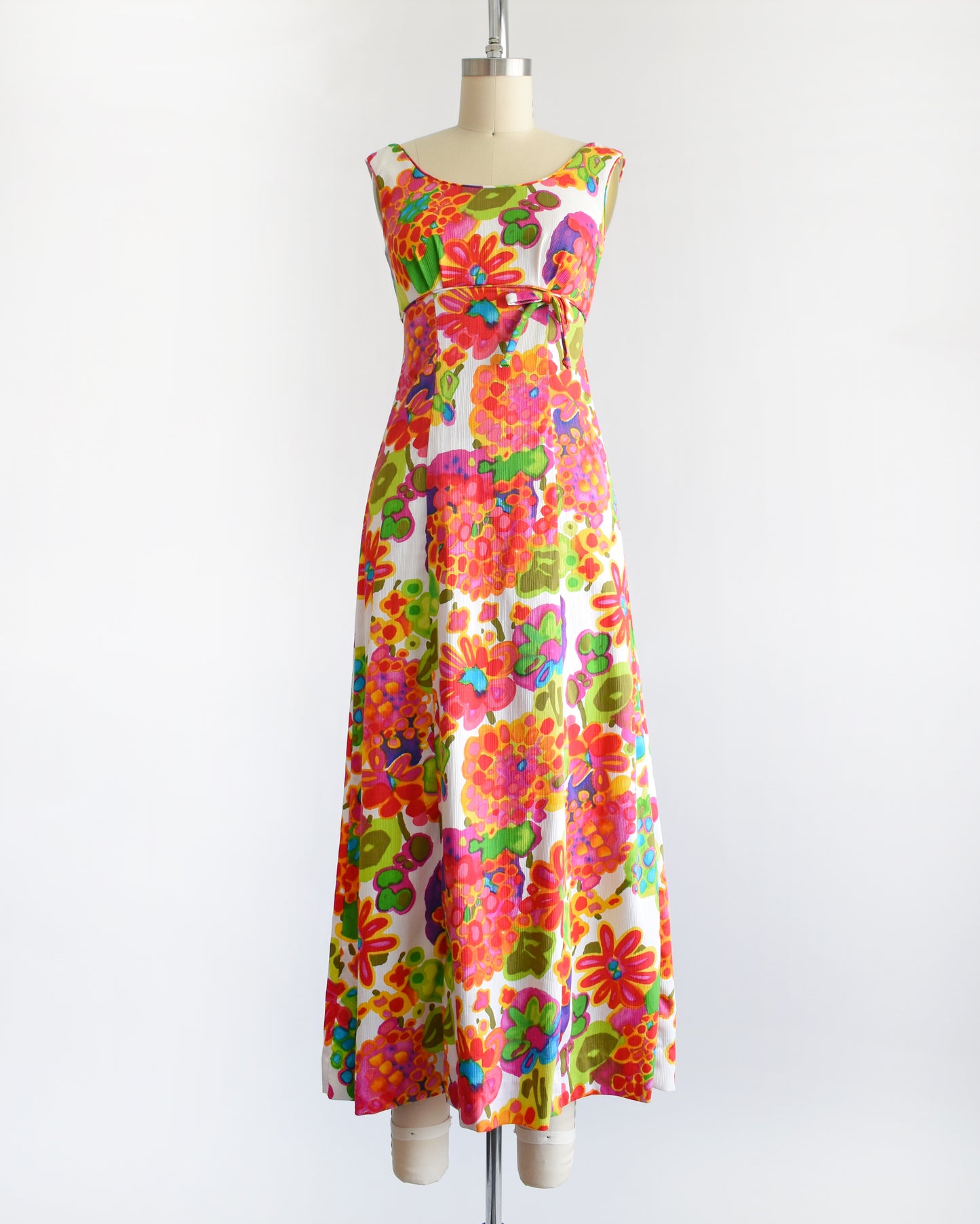 a vintage 1960s sleeveless white maxi dress with a vibrant, multicolored retro floral print, displayed on a cream-colored dress form against a plain white background.