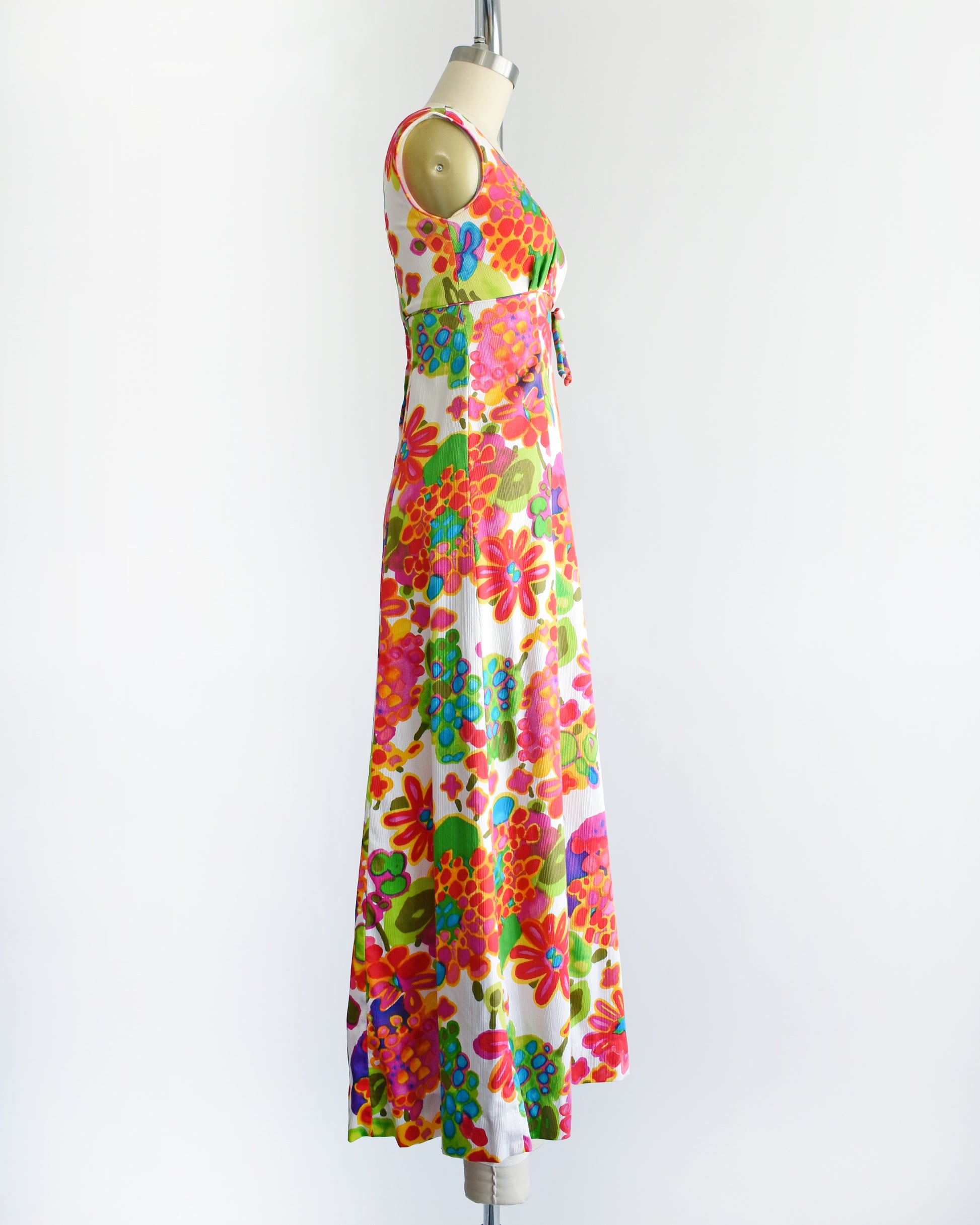 side view of a vintage 1960s sleeveless white maxi dress with a vibrant, multicolored retro floral print, displayed on a cream-colored dress form against a plain white background.