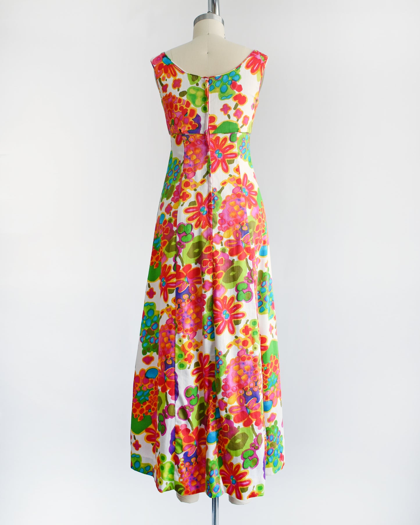 back view of a vintage 1960s sleeveless white maxi dress with a vibrant, multicolored retro floral print, displayed on a cream-colored dress form against a plain white background.