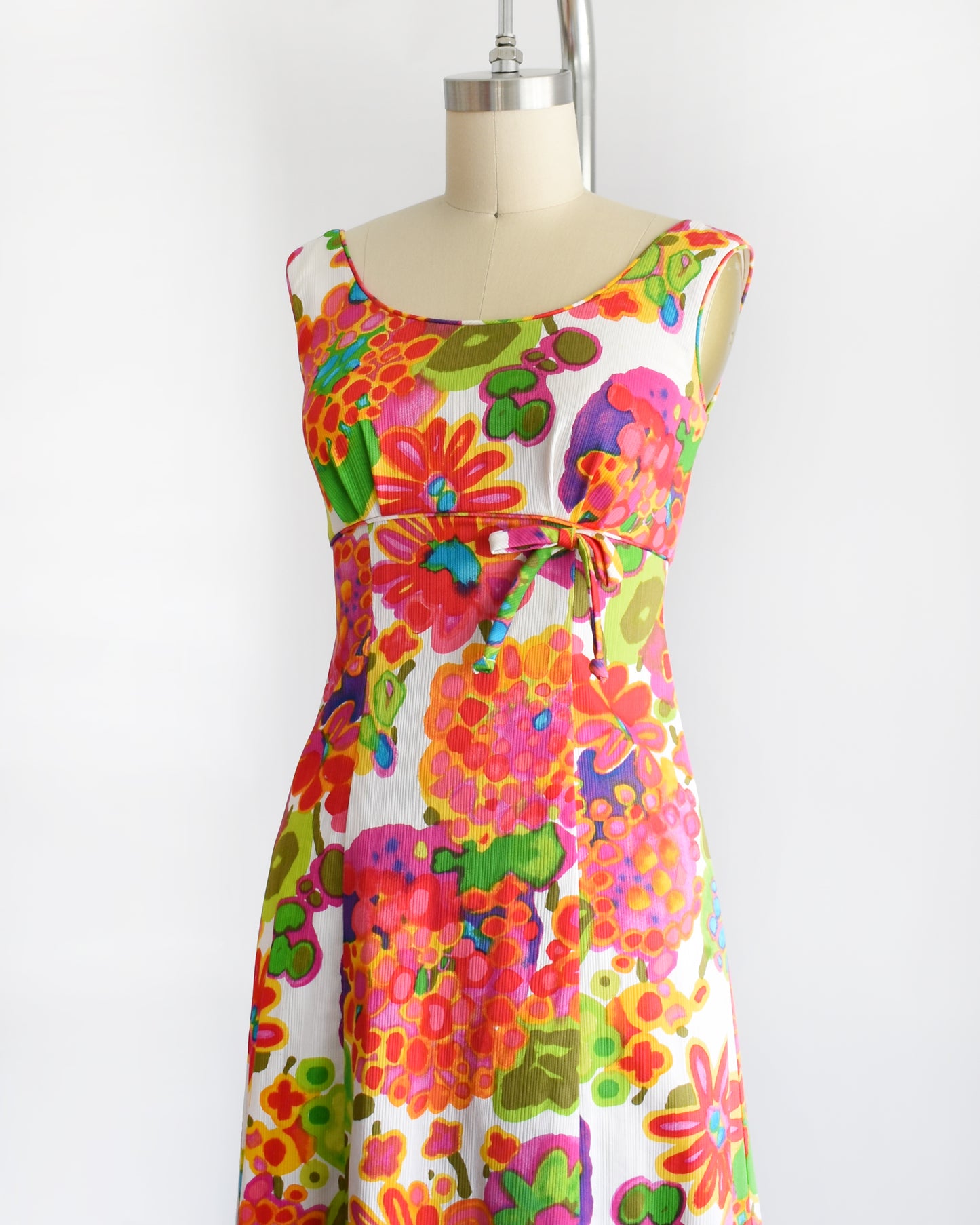side front view of a vintage 1960s sleeveless white maxi dress with a vibrant, multicolored retro floral print, displayed on a cream-colored dress form against a plain white background.