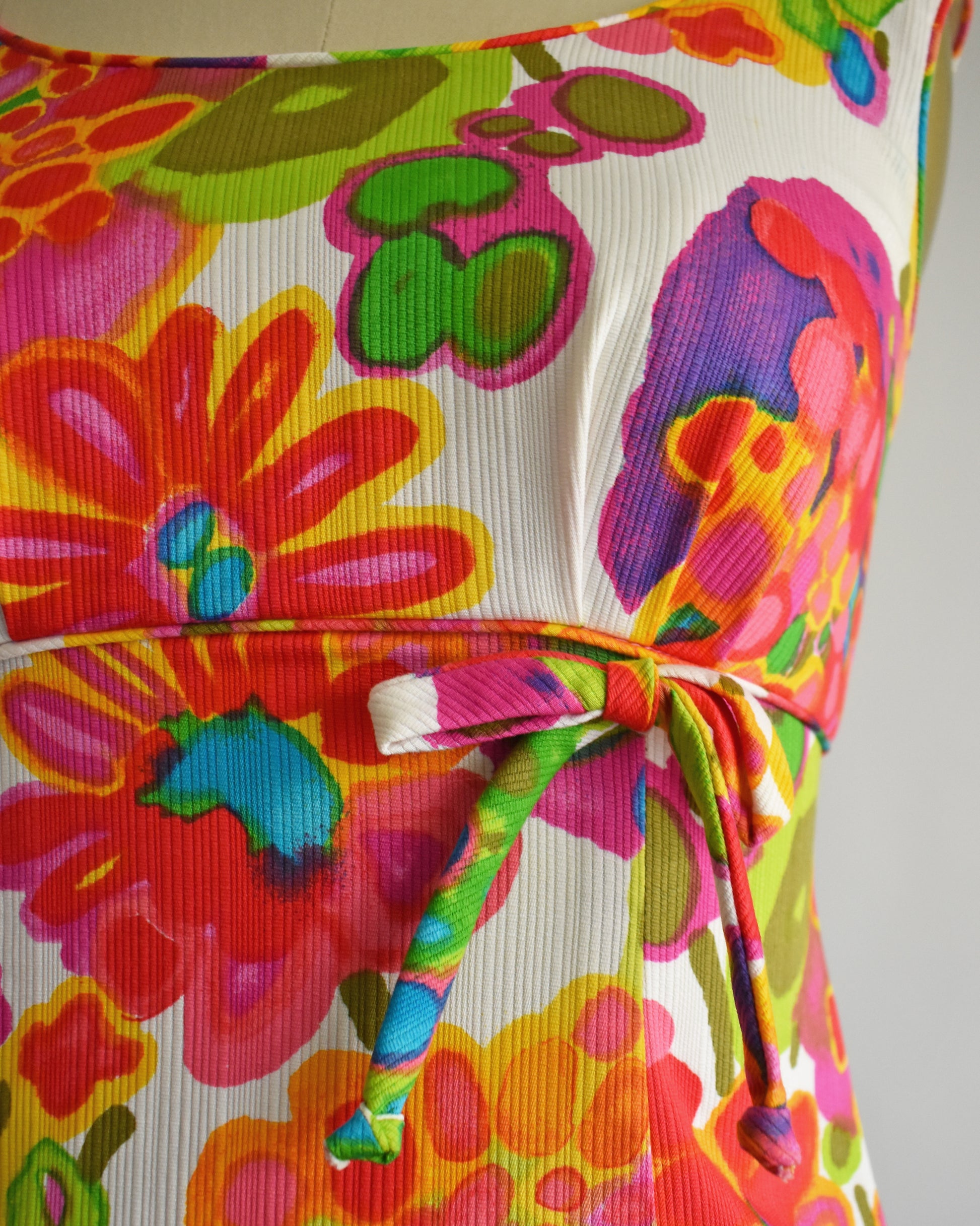 Close-up of a vintage 1960s floral dress with a tie waist, featuring bold pink, orange, green, and yellow flowers on a white background.