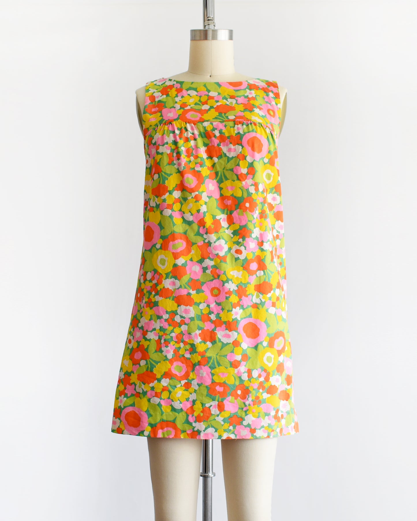 a vintage 1960s sleeveless A-line dress with a vibrant multicolor floral print, displayed on a cream-colored dress form mannequin against a plain white background.