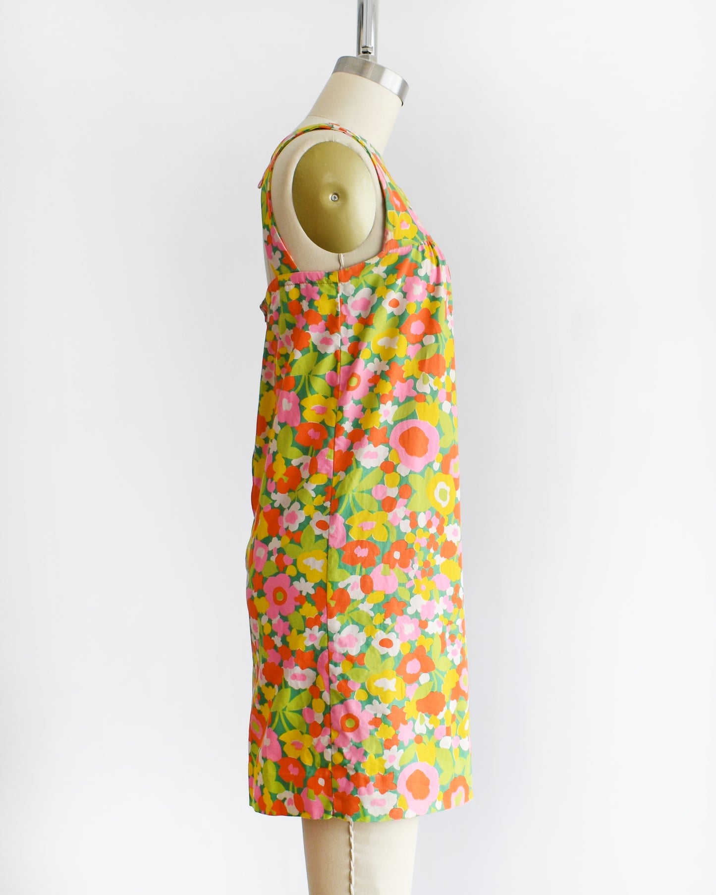 side view of a vintage 1960s sleeveless A-line dress with a vibrant multicolor floral print, displayed on a cream-colored dress form mannequin against a plain white background.