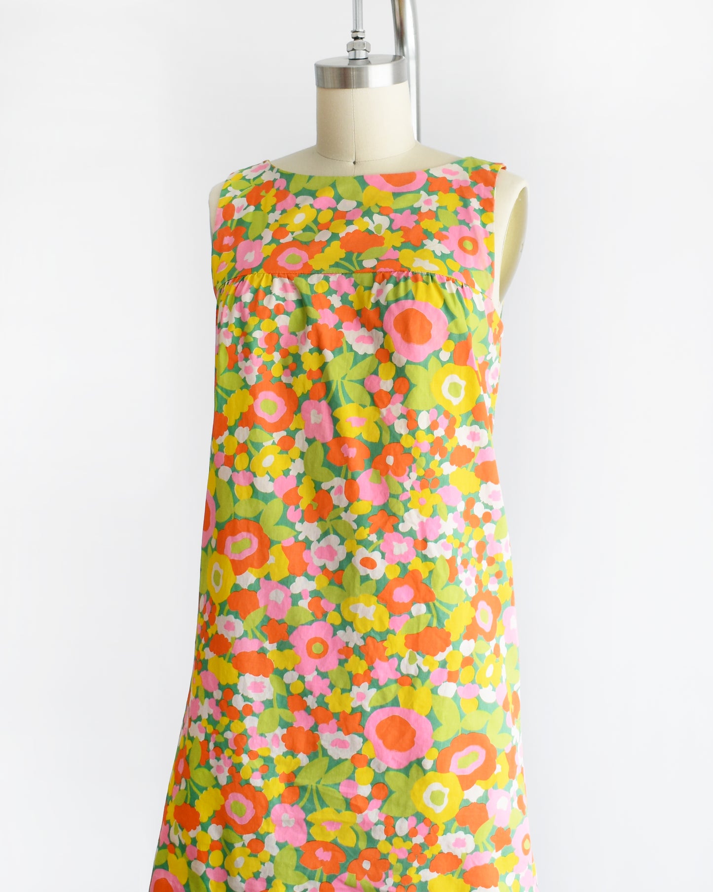 side front view of a vintage 1960s sleeveless A-line dress with a vibrant multicolor floral print, displayed on a cream-colored dress form mannequin against a plain white background.