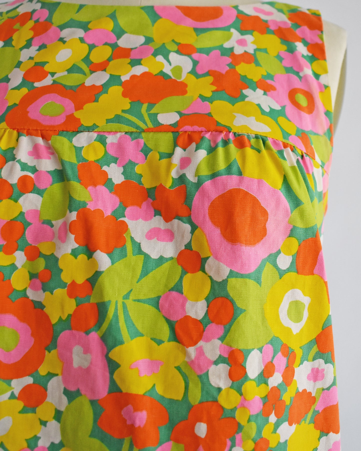 A close-up of a sleeveless vintge 1960s dress featuring a retro 1960s floral pattern with pink, orange, yellow, and green flowers on a teal background.