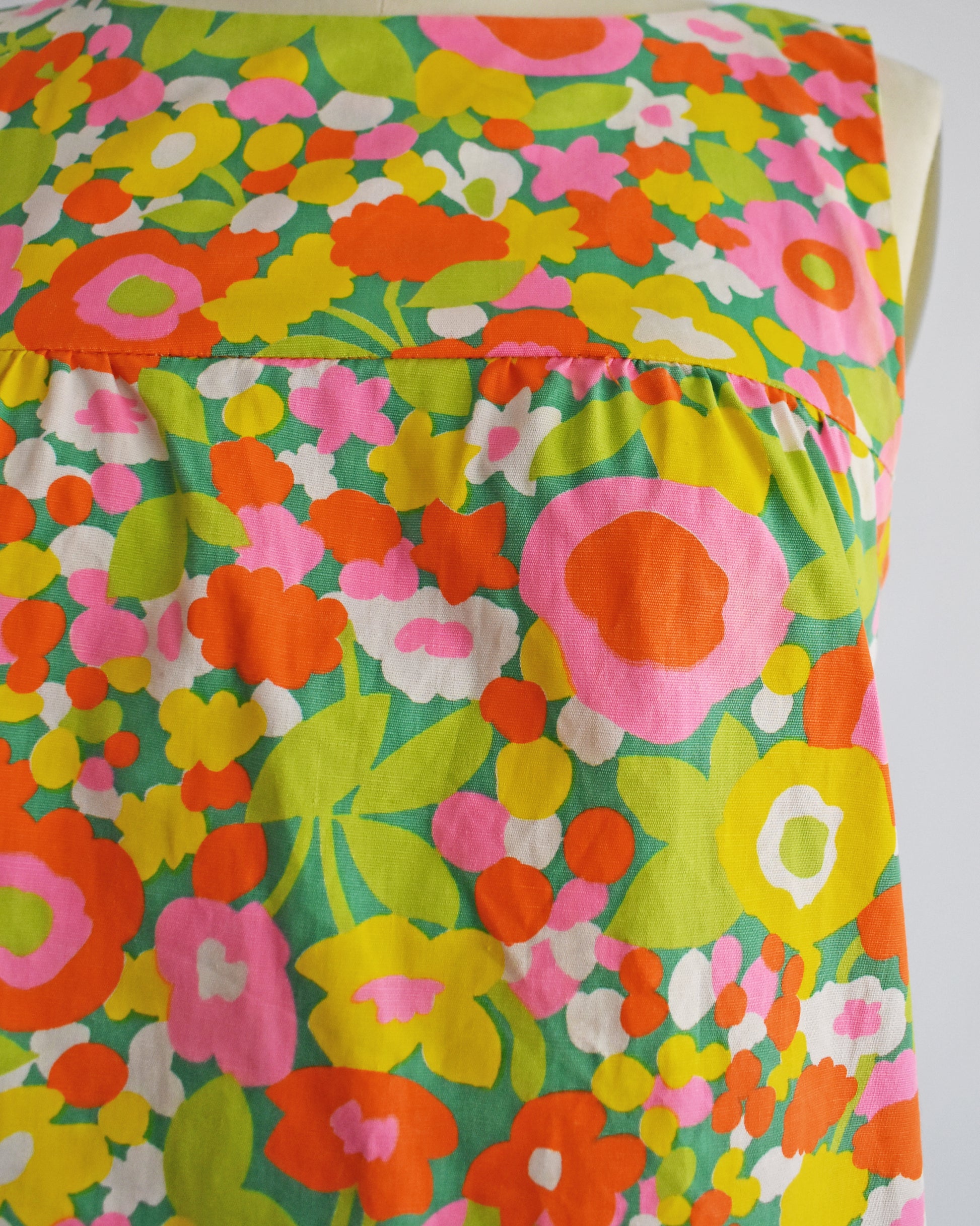 A close-up of a sleeveless vintge 1960s dress featuring a retro 1960s floral pattern with pink, orange, yellow, and green flowers on a teal background.