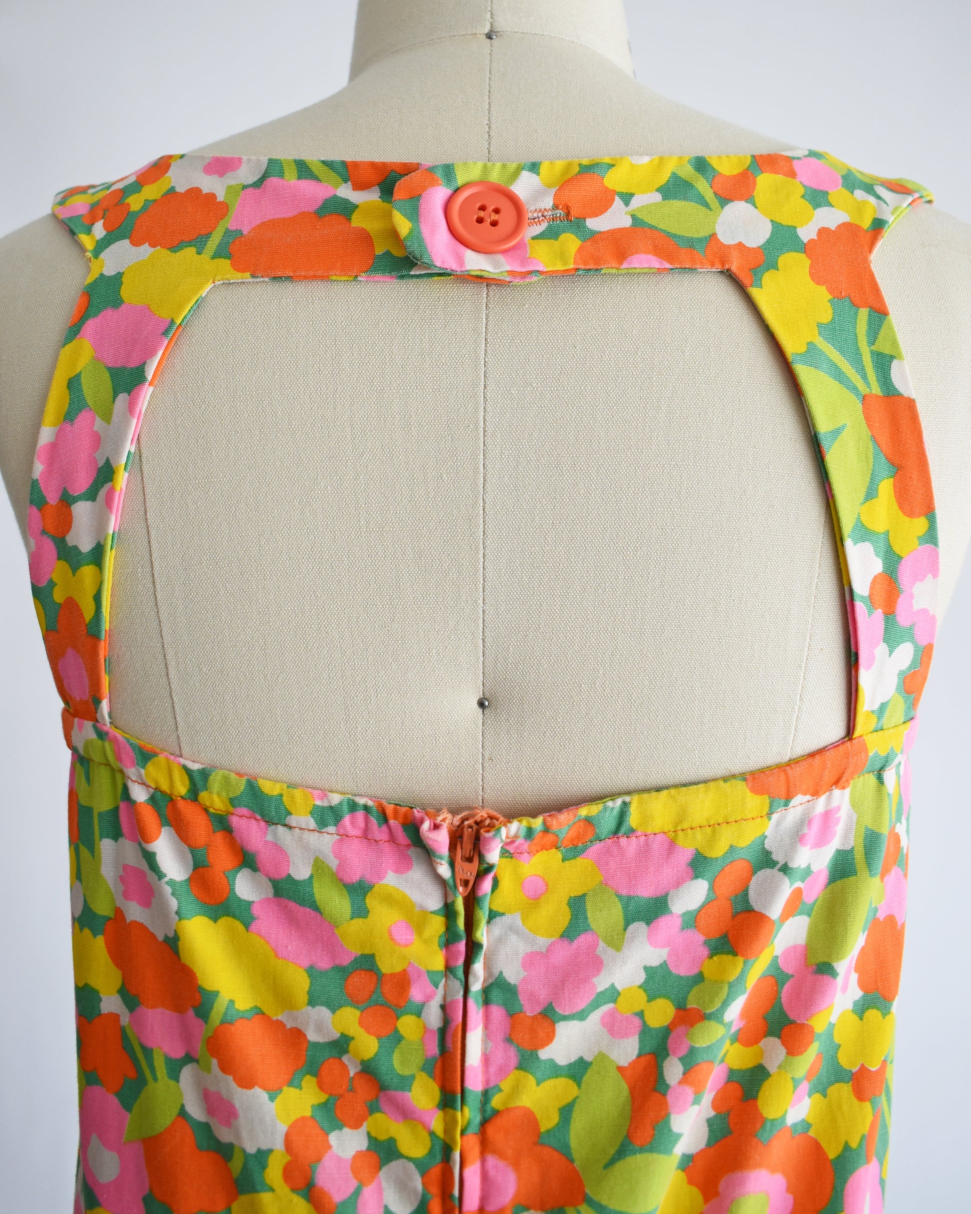 close up of the large cutout back on a vintage 1960s sleeveless A-line dress with a vibrant multicolor floral print, displayed on a cream-colored dress form mannequin against a plain white background.