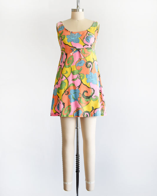 A vintage 1960s sleeveless mini dress with a vibrant floral pattern in pink, yellow, green, and blue, displayed on a beige dress form mannequin against a white background.