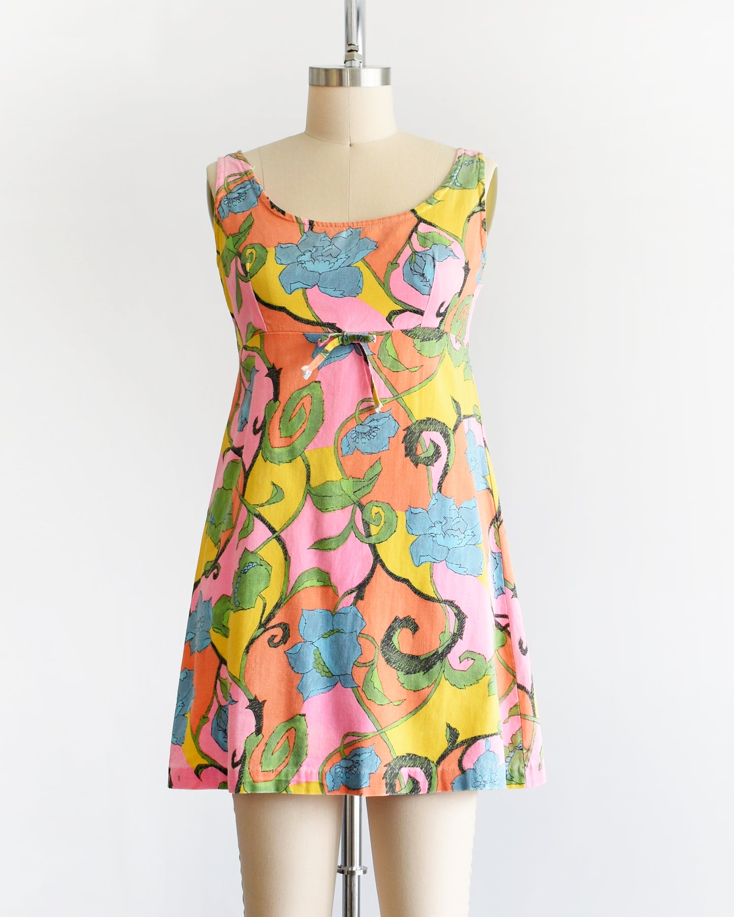 A vintage 1960s sleeveless mini dress with a vibrant floral pattern in pink, yellow, green, and blue, displayed on a beige dress form mannequin against a white background.