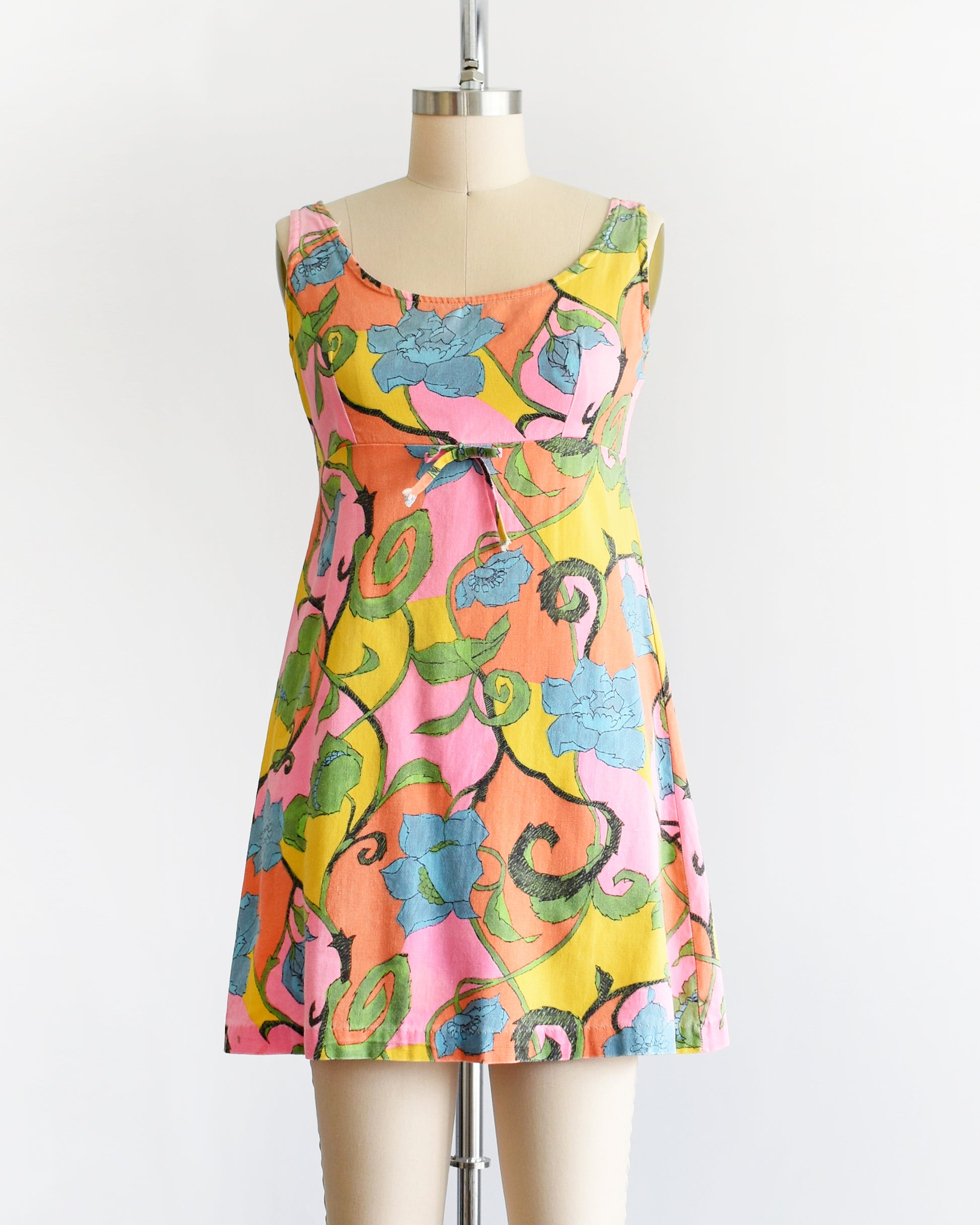 A vintage 1960s sleeveless mini dress with a vibrant floral pattern in pink, yellow, green, and blue, displayed on a beige dress form mannequin against a white background.