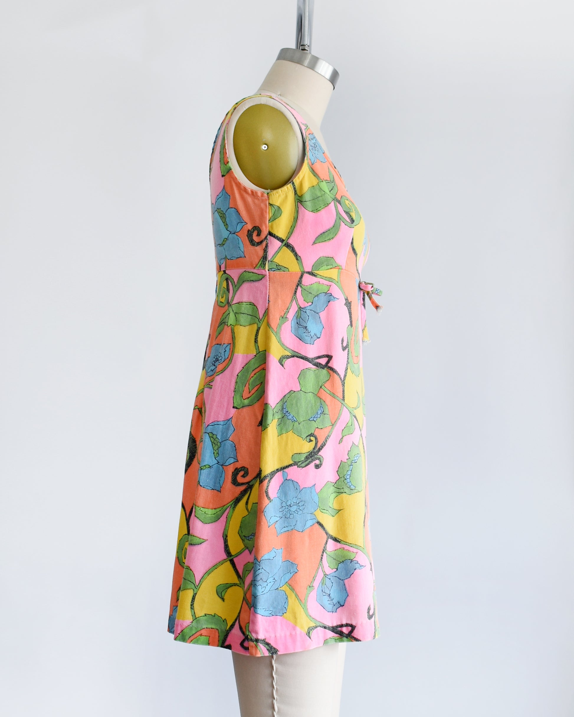 a side view of a vintage 1960s sleeveless mini dress with a vibrant floral pattern in pink, yellow, green, and blue, displayed on a beige dress form mannequin against a white background.