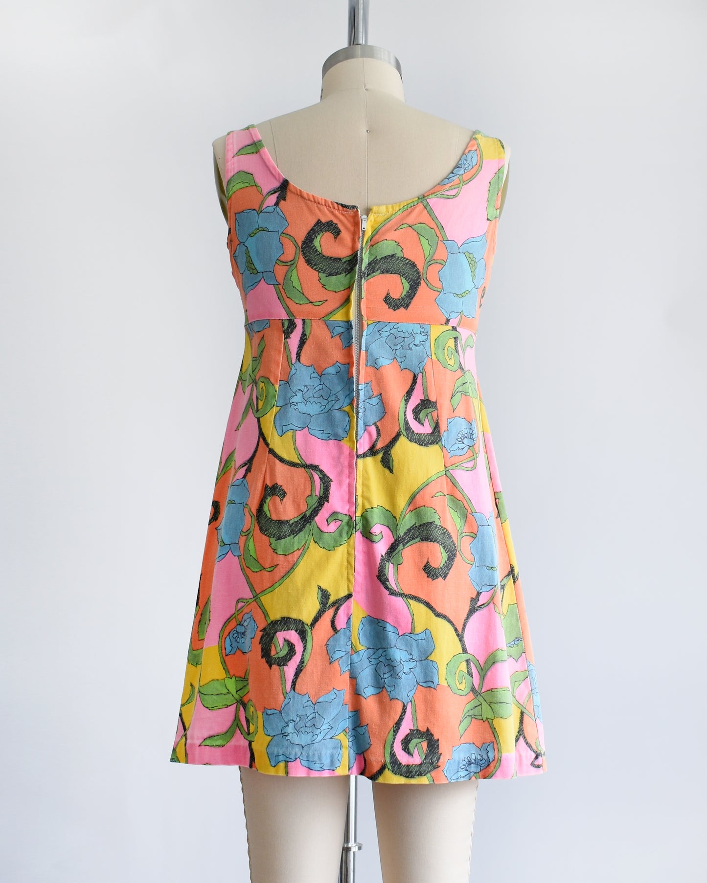 back view of  a vintage 1960s sleeveless mini dress with a vibrant floral pattern in pink, yellow, green, and blue, displayed on a beige dress form mannequin against a white background.