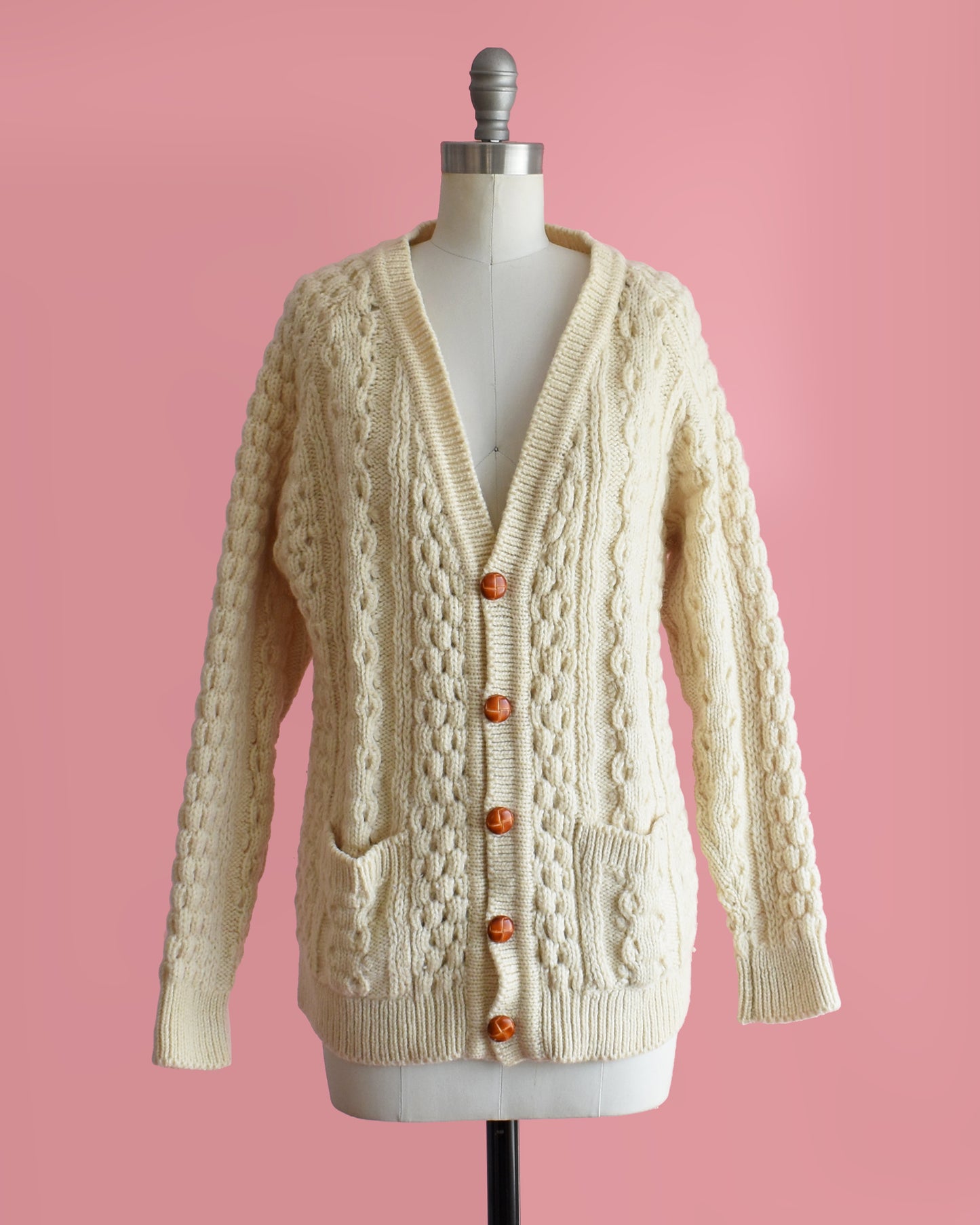 a vintage 1980s cream-colored cable knit cardigan on a mannequin against a pink background