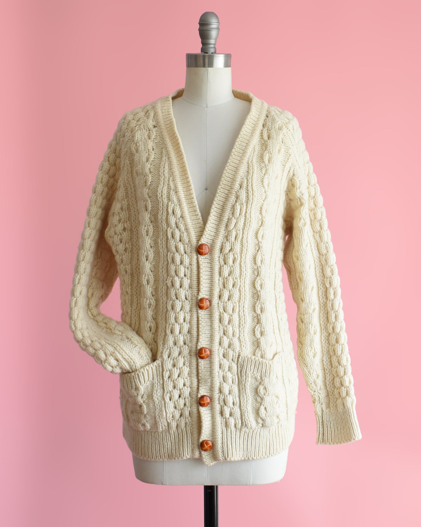 a vintage 1980s cream-colored cable knit cardigan on a mannequin against a pink background