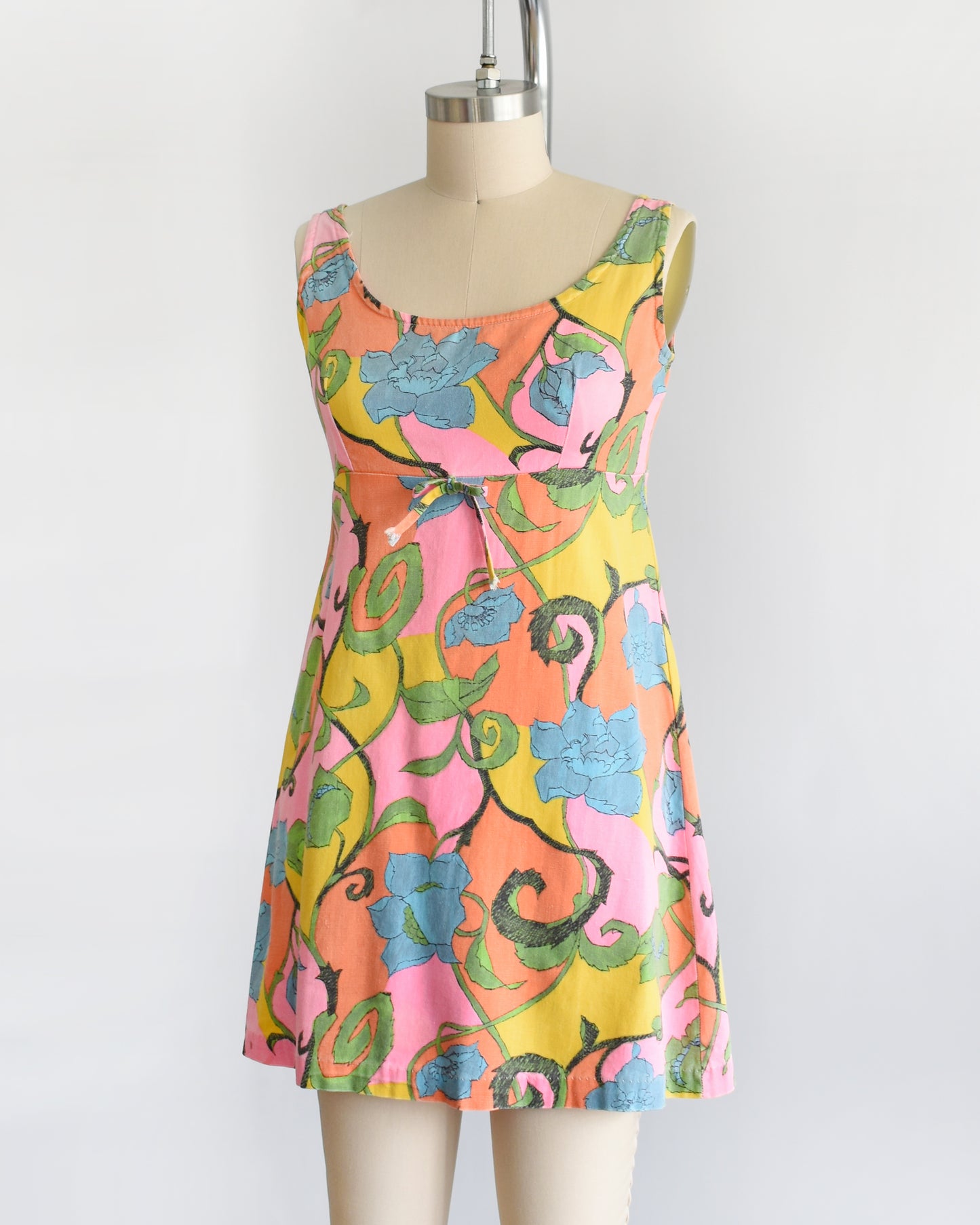 side front view of  a vintage 1960s sleeveless mini dress with a vibrant floral pattern in pink, yellow, green, and blue, displayed on a beige dress form mannequin against a white background.