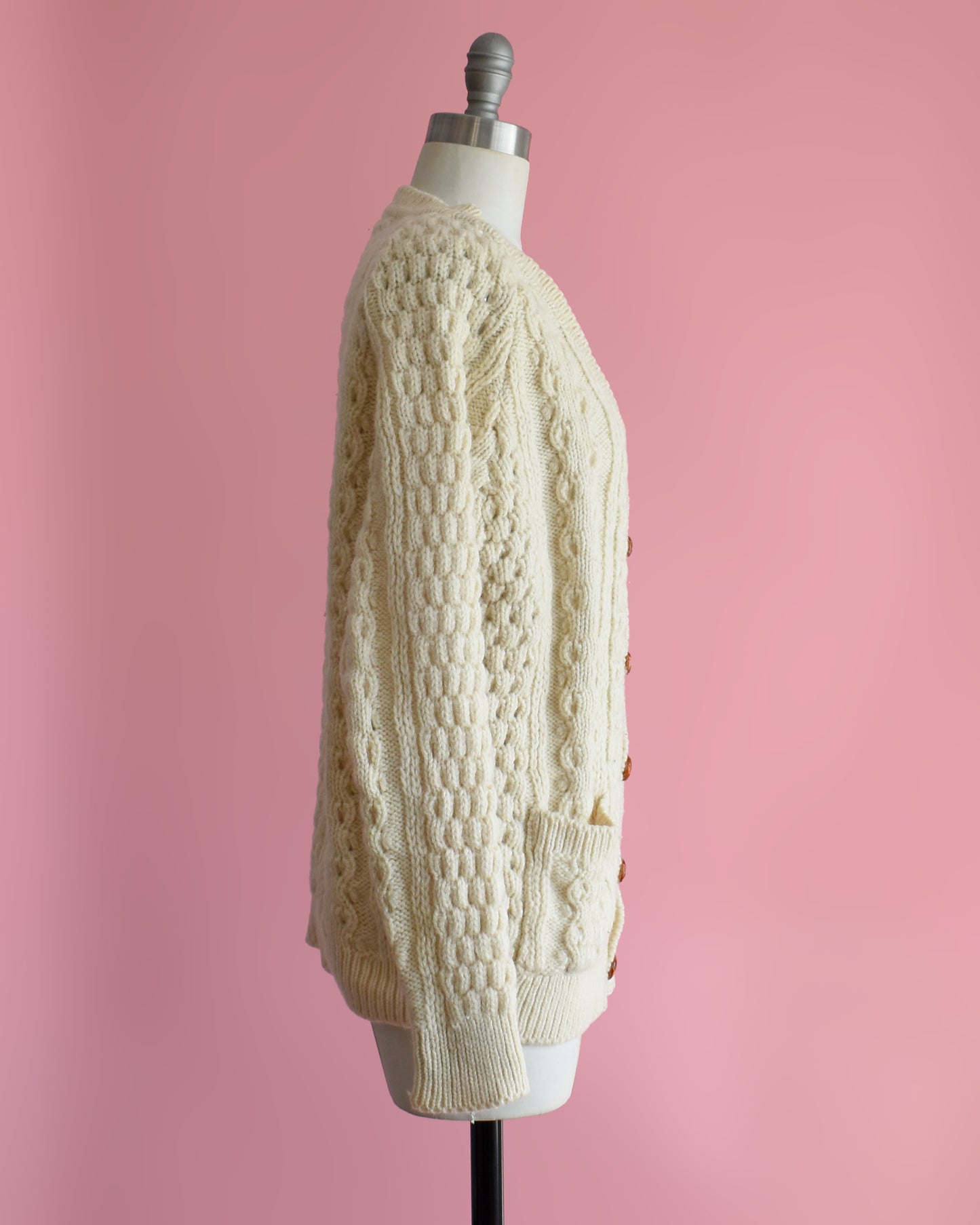 side view of a vintage 1980s cream-colored cable knit cardigan on a mannequin against a pink background