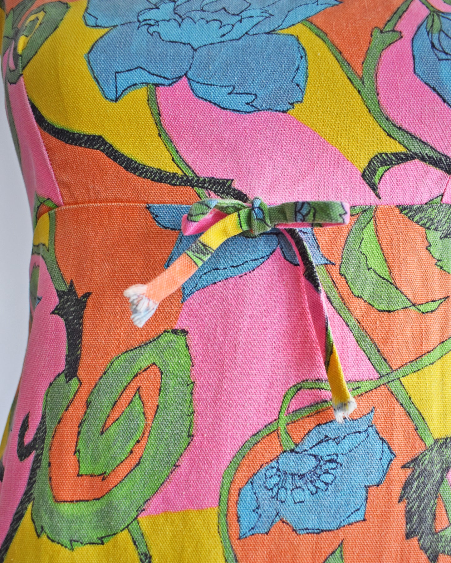 close up of the decorative bow on  a vintage 1960s sleeveless mini dress with a vibrant floral pattern in pink, yellow, green, and blue, displayed on a beige dress form mannequin against a white background.
