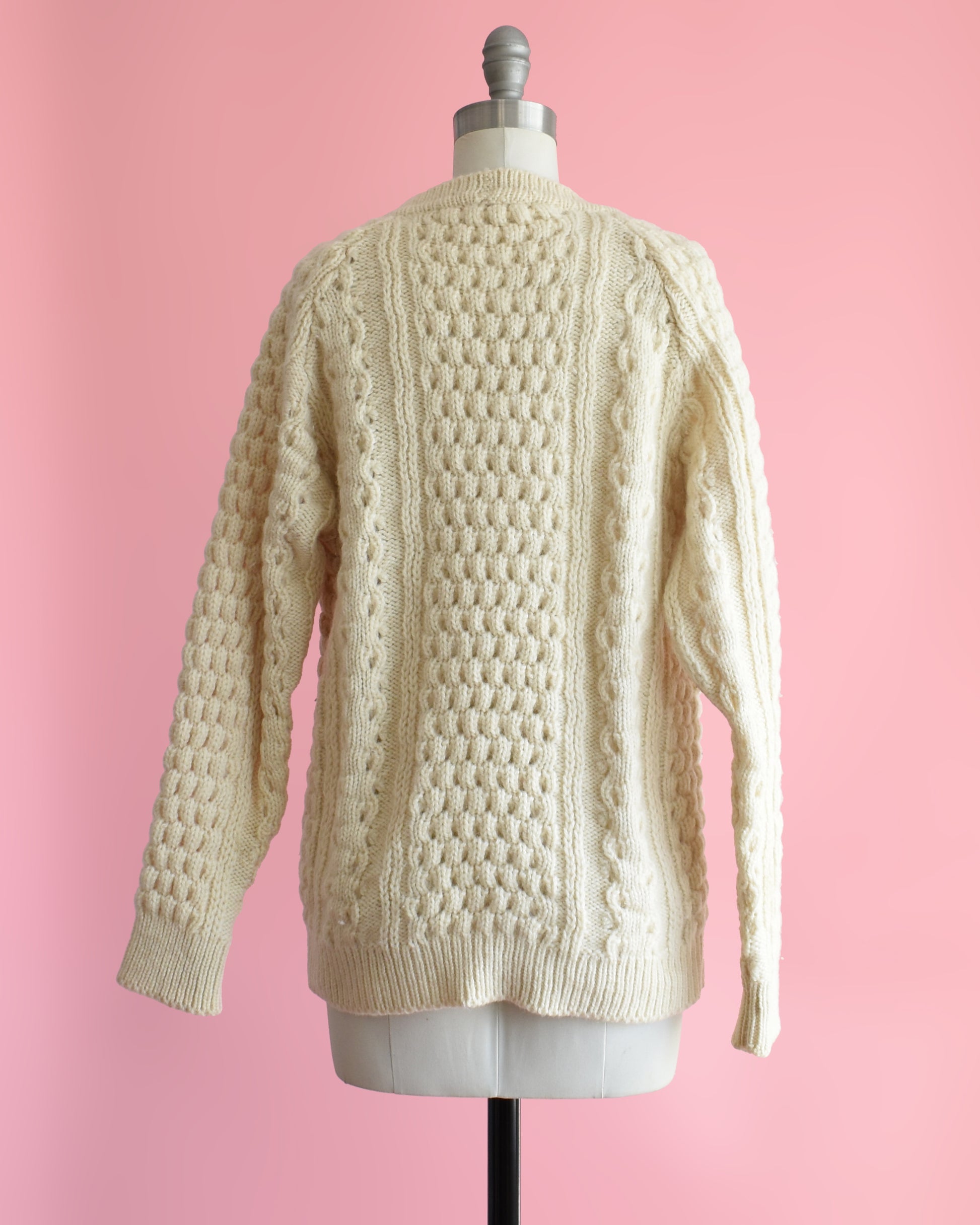 back view of a vintage 1980s cream-colored cable knit cardigan on a mannequin against a pink background