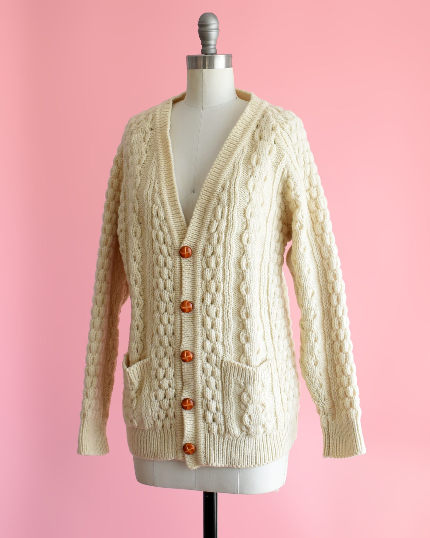side front view of a vintage 1980s cream-colored cable knit cardigan on a mannequin against a pink background