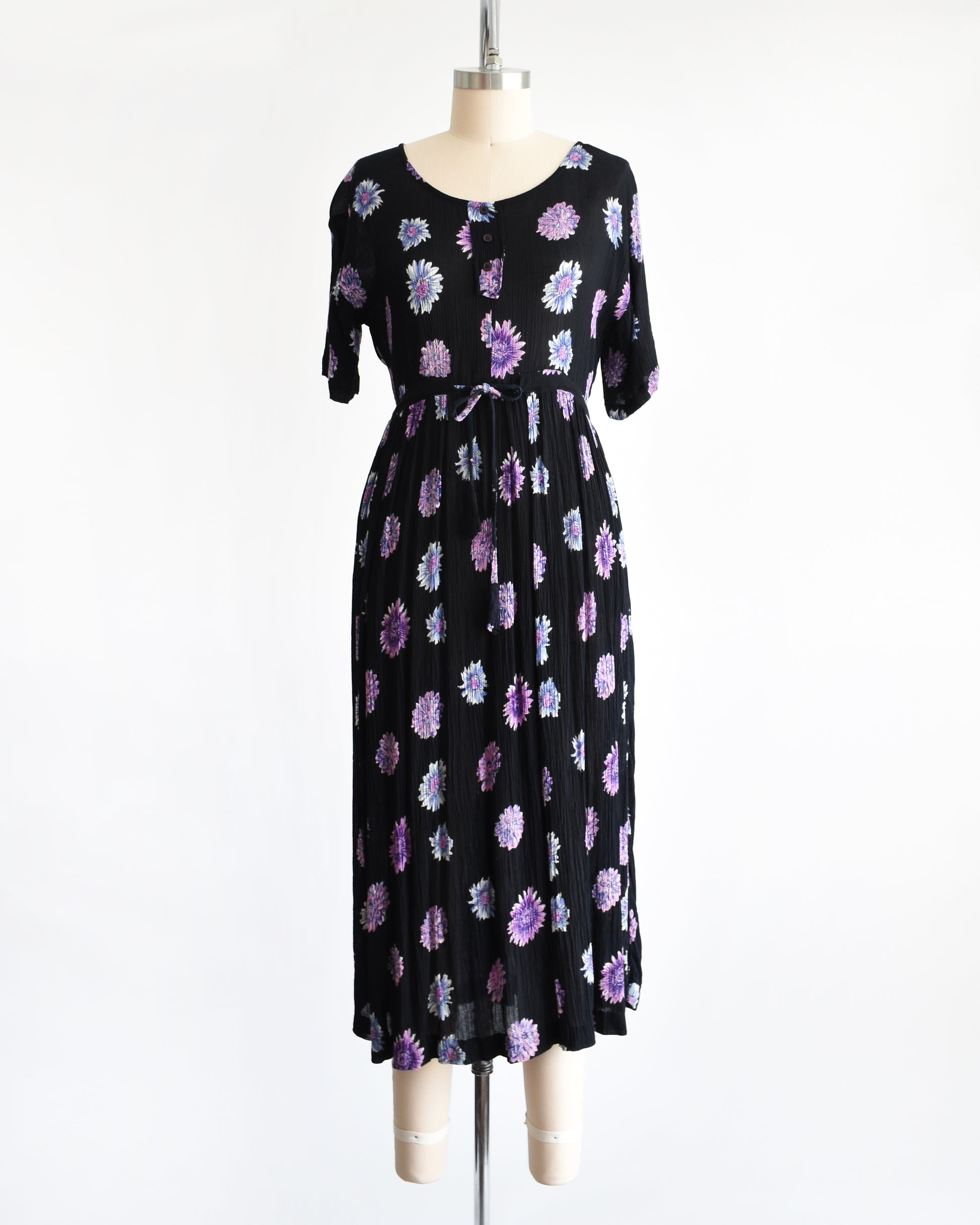 a vintage 1970s black midi dress with a purple and blue floral pattern, displayed on a mannequin against a plain white background.