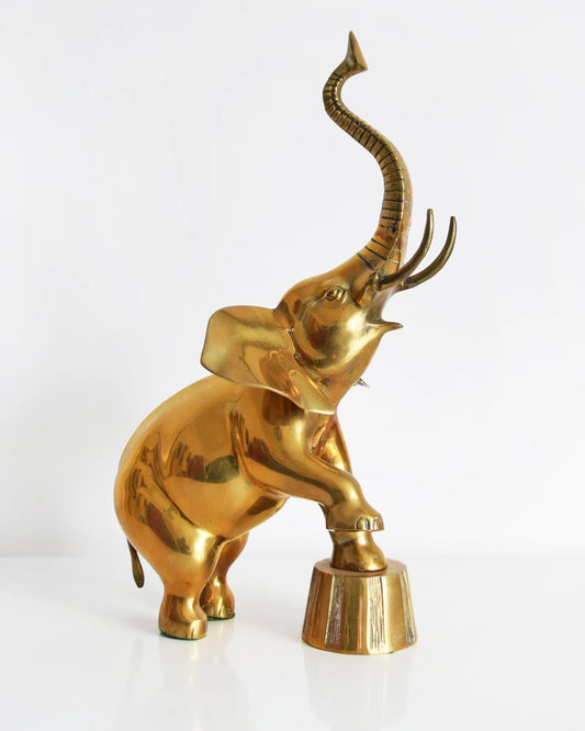 A large brass elephant that is balancing one of its front legs on a a platform and proudly displaying its trunk up for good luck.