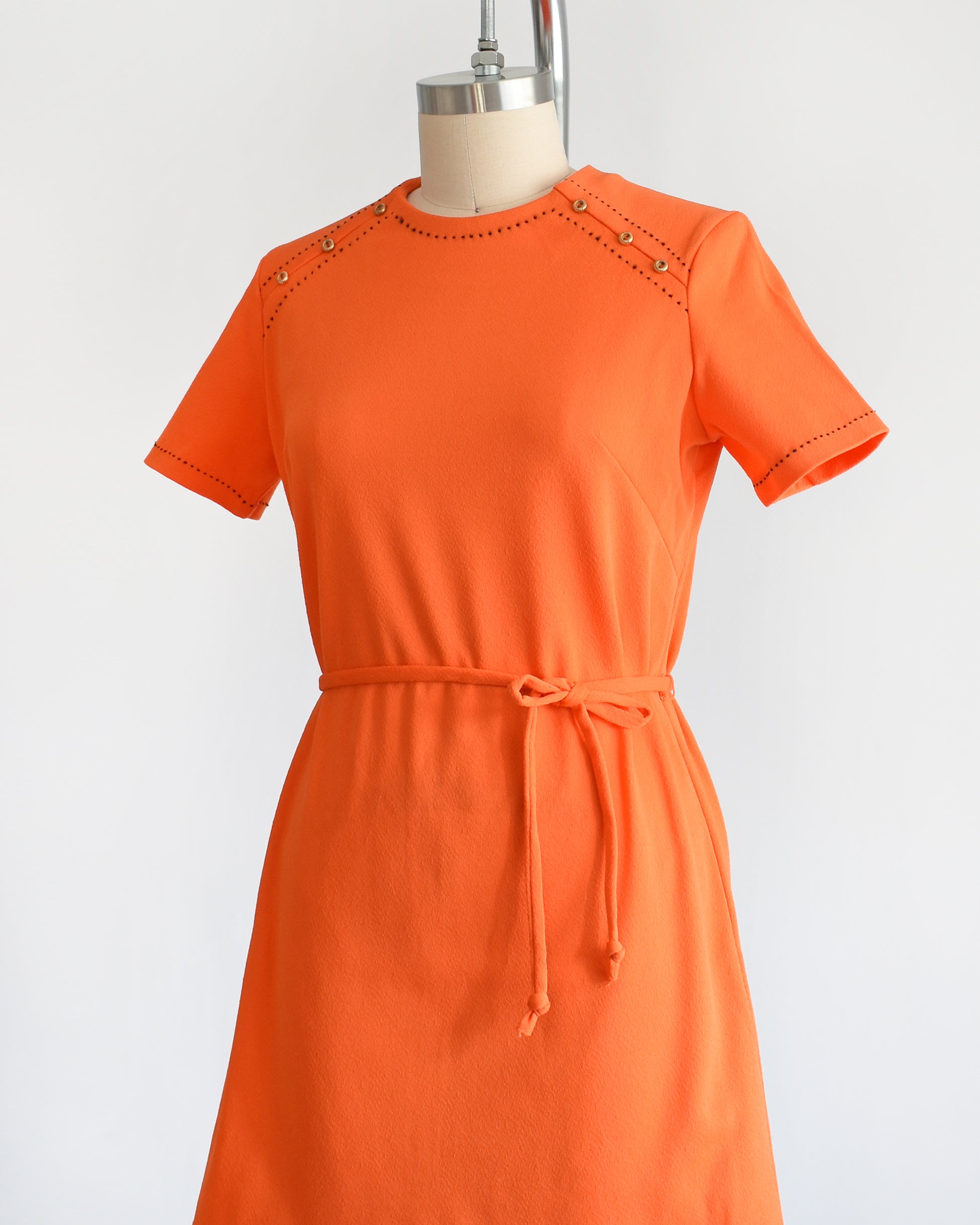 side front view of a vintage 1960s orange mod dress with black stitch trim on the front collar and shoulders, along with decorative gold buttons on the shoulder.