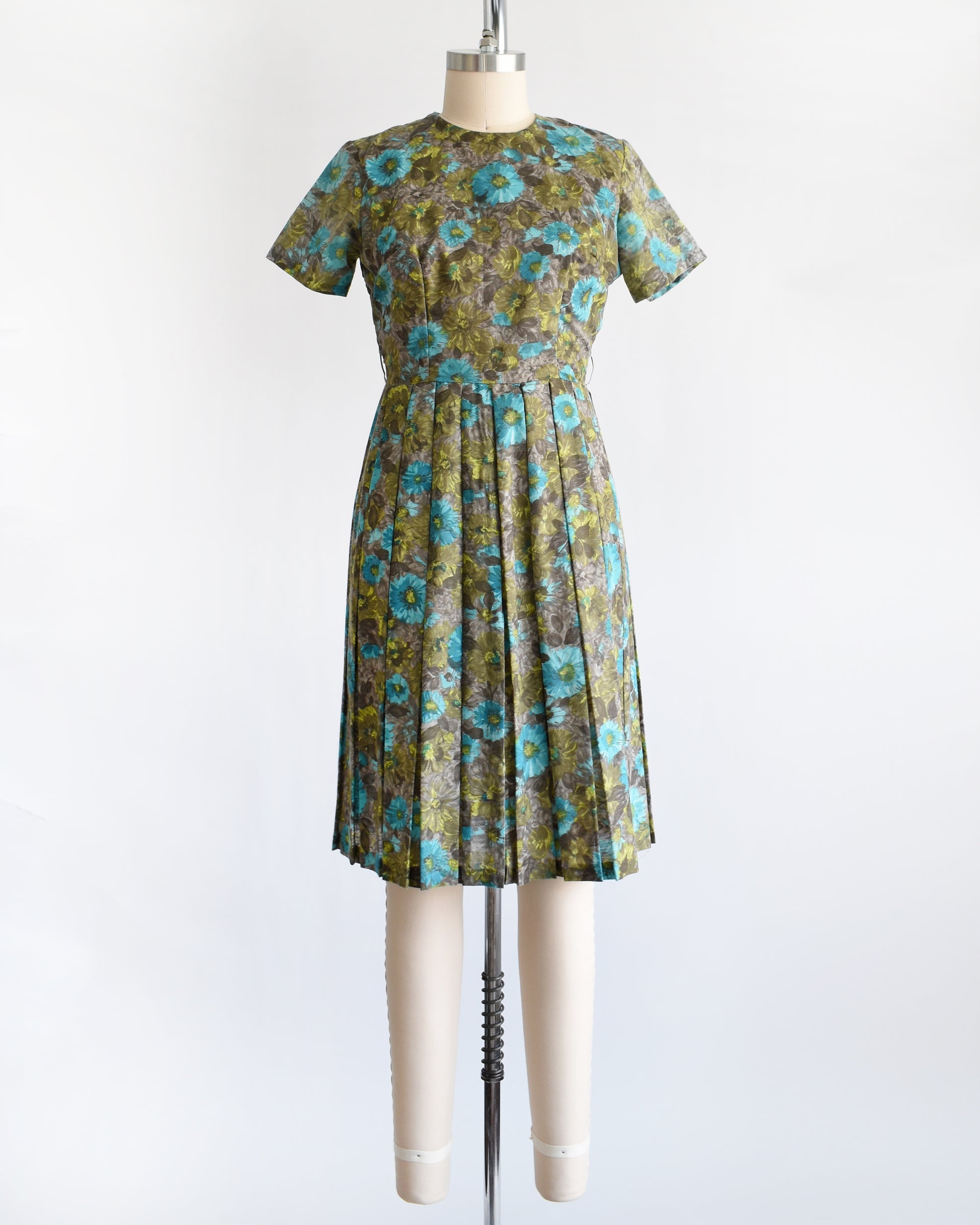 A vintage 1950s short-sleeve knee-length dress with a pleated skirt and gathered bodice, featuring a blue and green floral pattern on a brown base, displayed on a cream dress form against a white background.