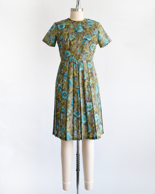 A vintage 1950s short-sleeve knee-length dress with a pleated skirt and gathered bodice, featuring a blue and green floral pattern on a brown base, displayed on a cream dress form against a white background.