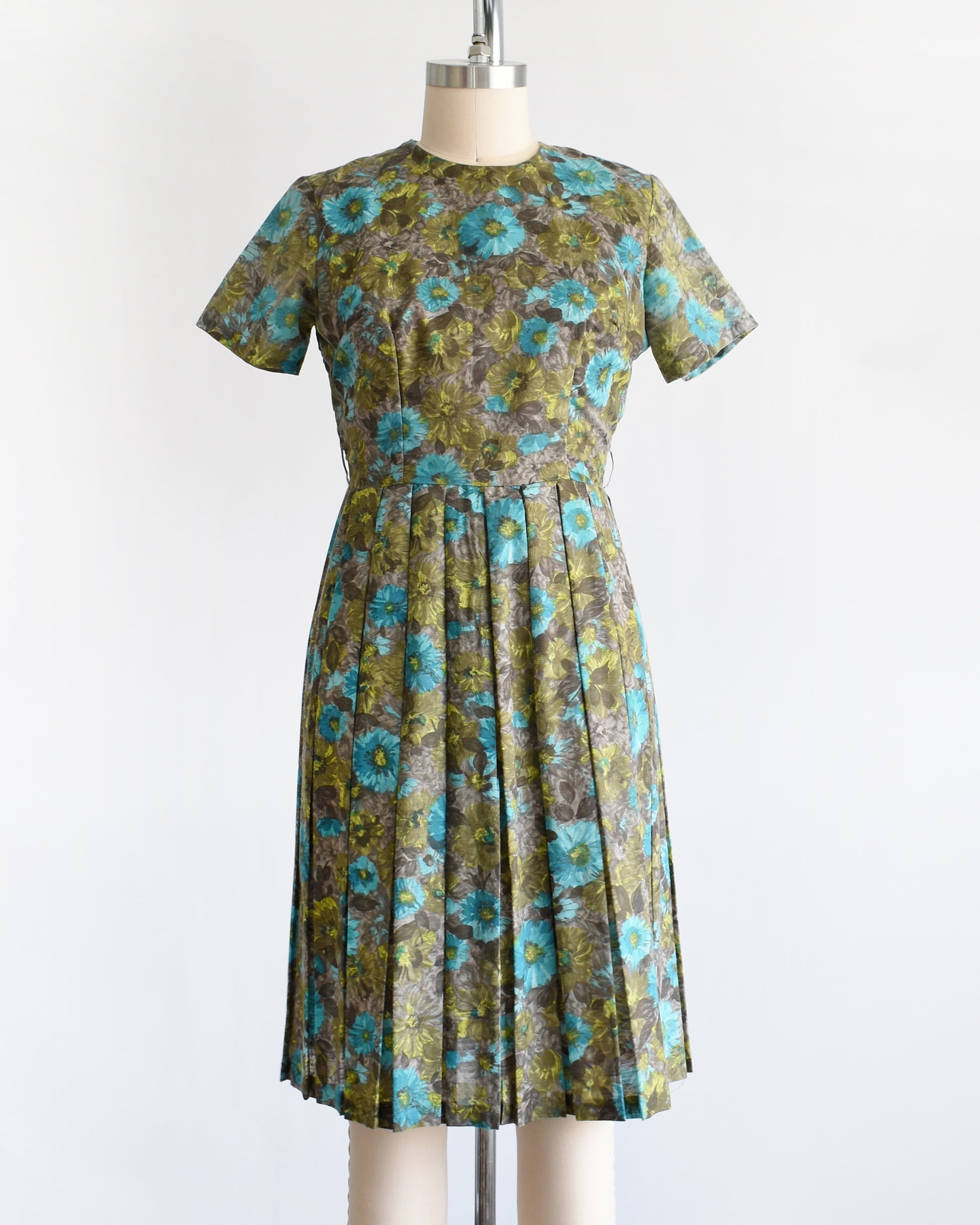 A vintage 1950s short-sleeve knee-length dress with a pleated skirt and gathered bodice, featuring a blue and green floral pattern on a brown base, displayed on a cream dress form against a white background.