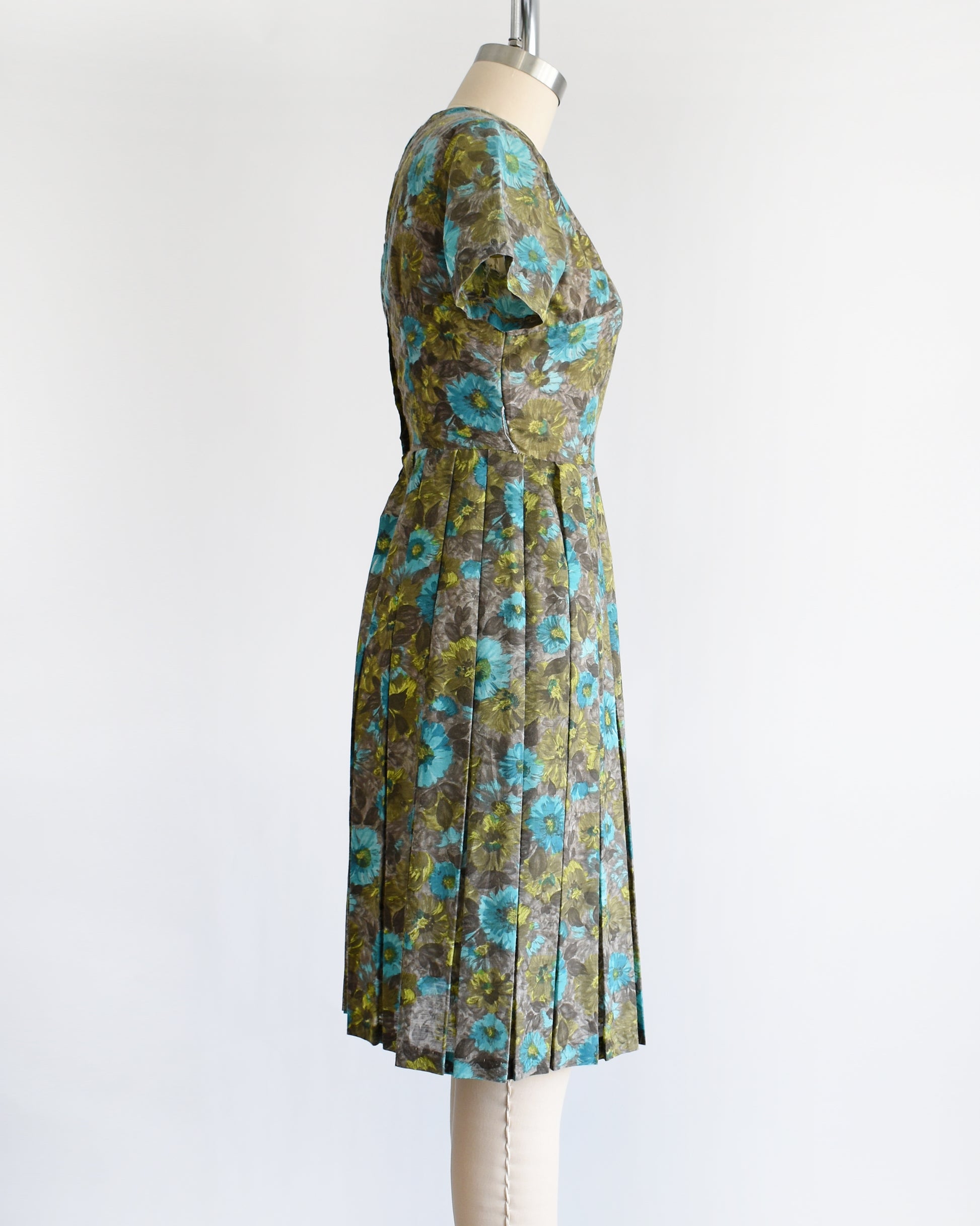 side view of a vintage 1950s short-sleeve knee-length dress with a pleated skirt and gathered bodice, featuring a blue and green floral pattern on a brown base, displayed on a cream dress form against a white background.