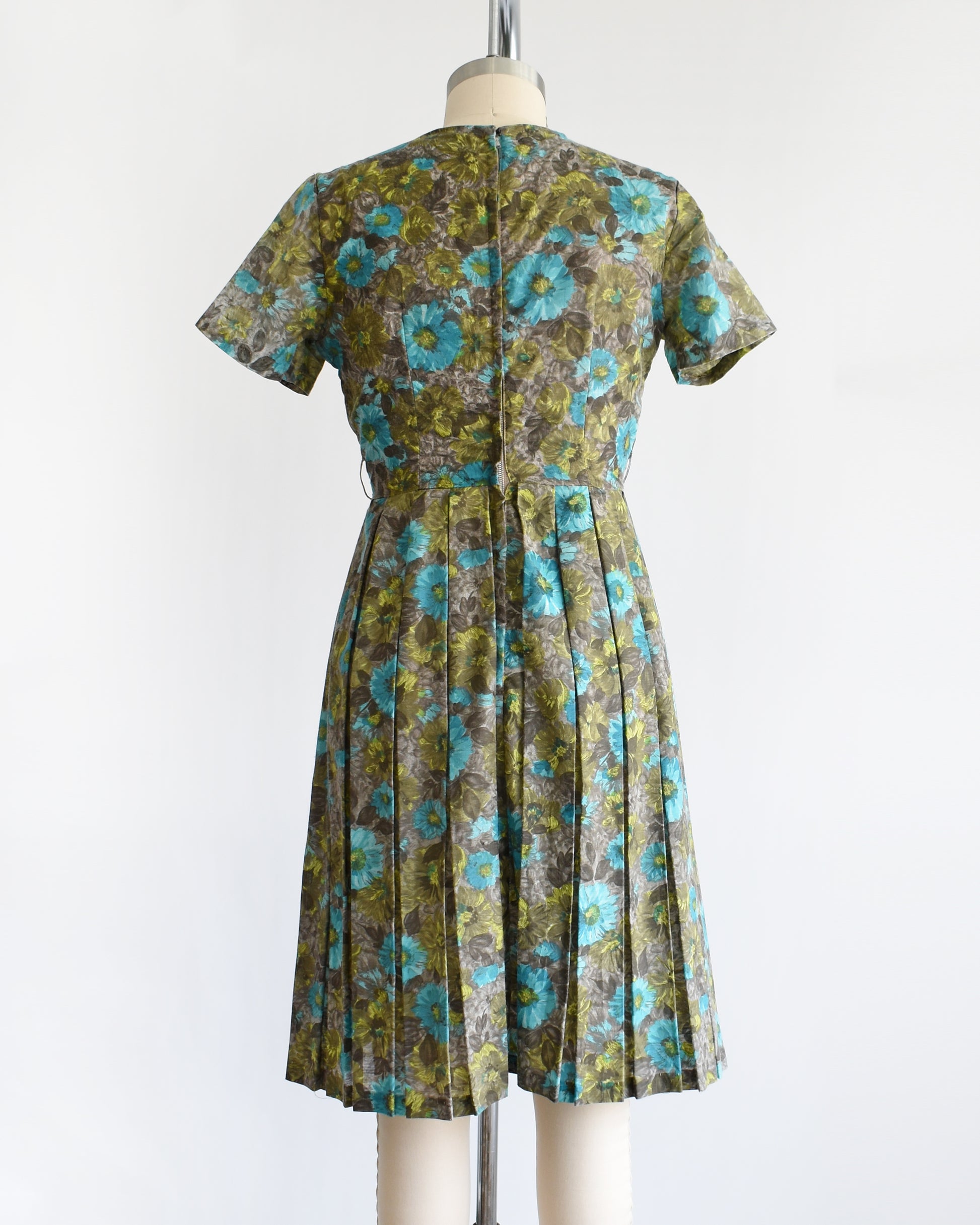 back view of a vintage 1950s short-sleeve knee-length dress with a pleated skirt and gathered bodice, featuring a blue and green floral pattern on a brown base, displayed on a cream dress form against a white background.