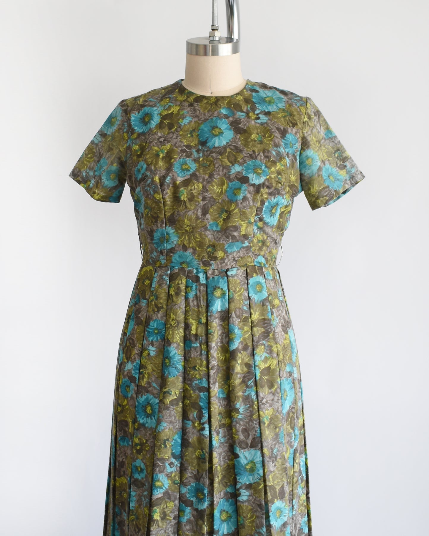 side front view of a vintage 1950s short-sleeve knee-length dress with a pleated skirt and gathered bodice, featuring a blue and green floral pattern on a brown base, displayed on a cream dress form against a white background.