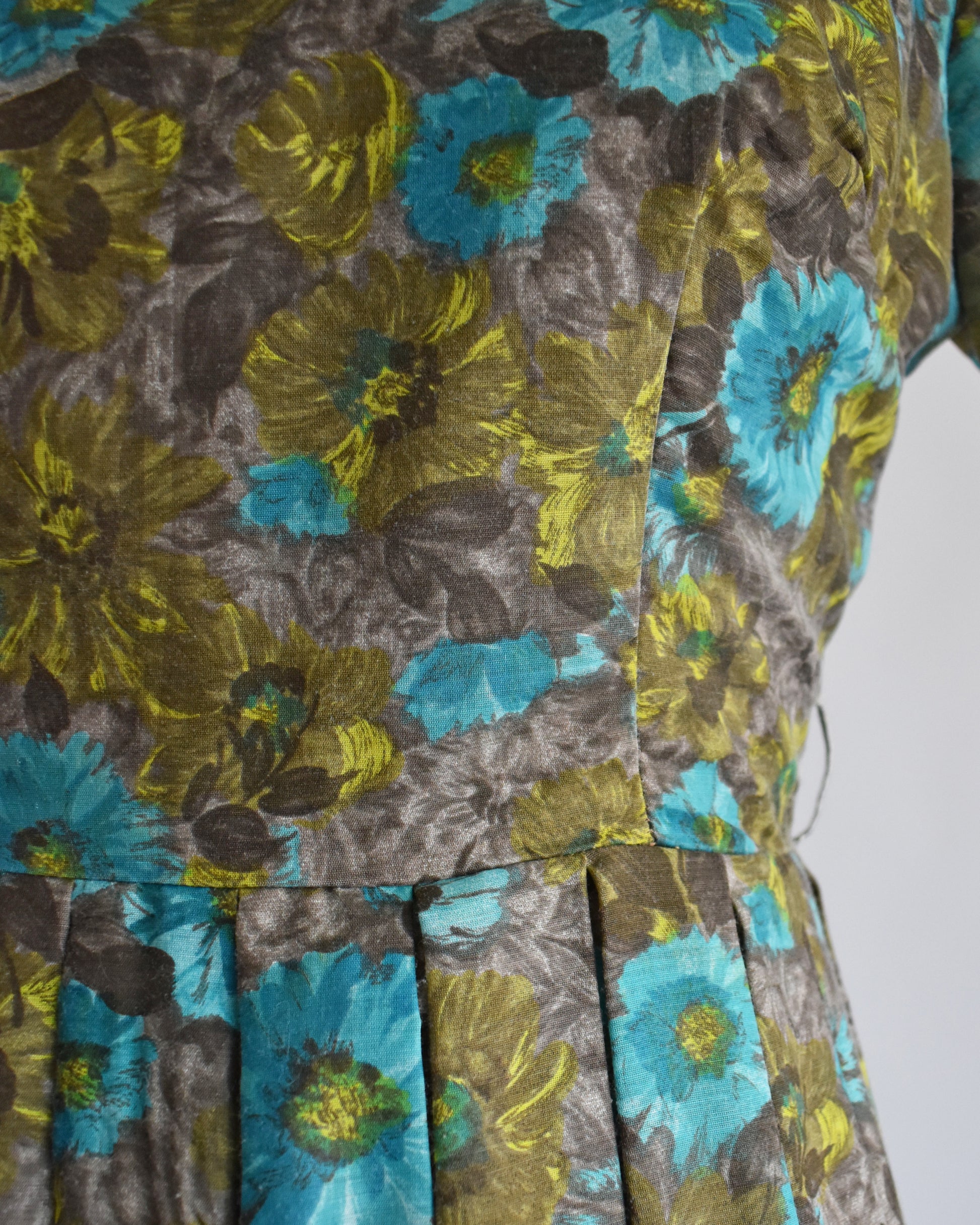 Close-up of a floral dress fabric with large yellow-green and turquoise flowers on a gray background, showing the pleated waistband detail.