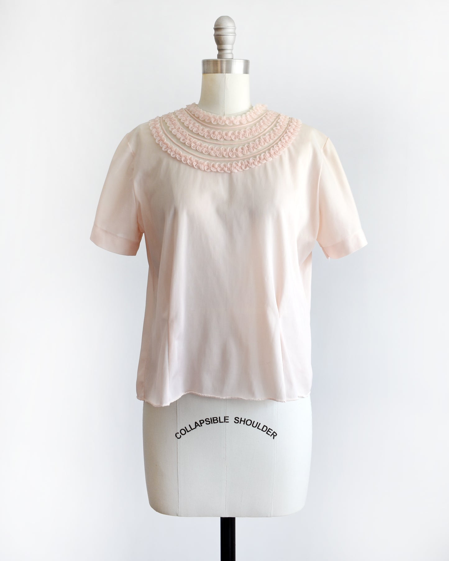 A vintage 1960s pale blush pink short-sleeve blouse with a decorative layered ruffle collar, displayed on a white mannequin dress form against a white background.