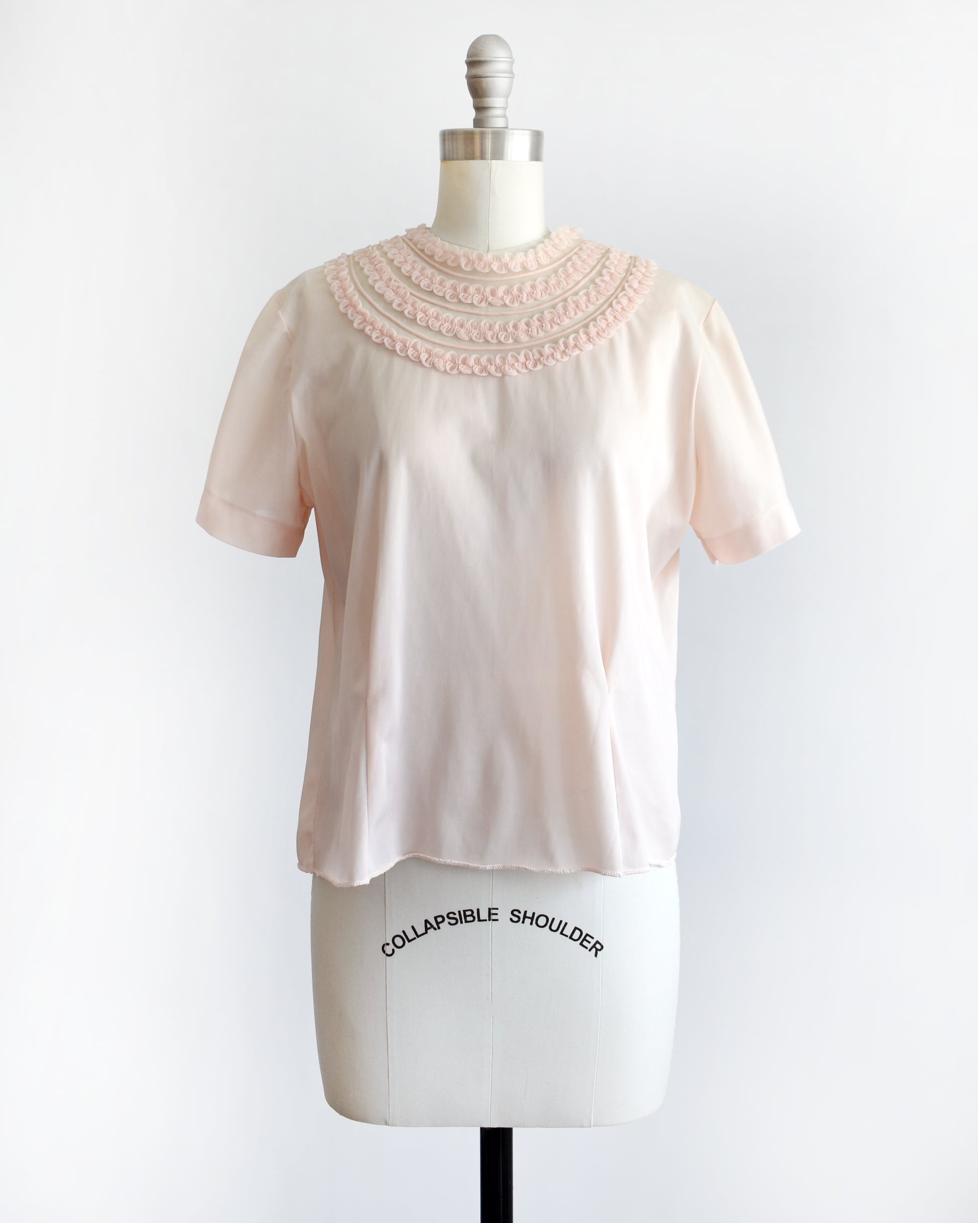 A vintage 1960s pale blush pink short-sleeve blouse with a decorative layered ruffle collar, displayed on a white mannequin dress form against a white background.