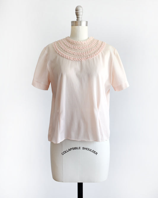A vintage 1960s pale blush pink short-sleeve blouse with a decorative layered ruffle collar, displayed on a white mannequin dress form against a white background.